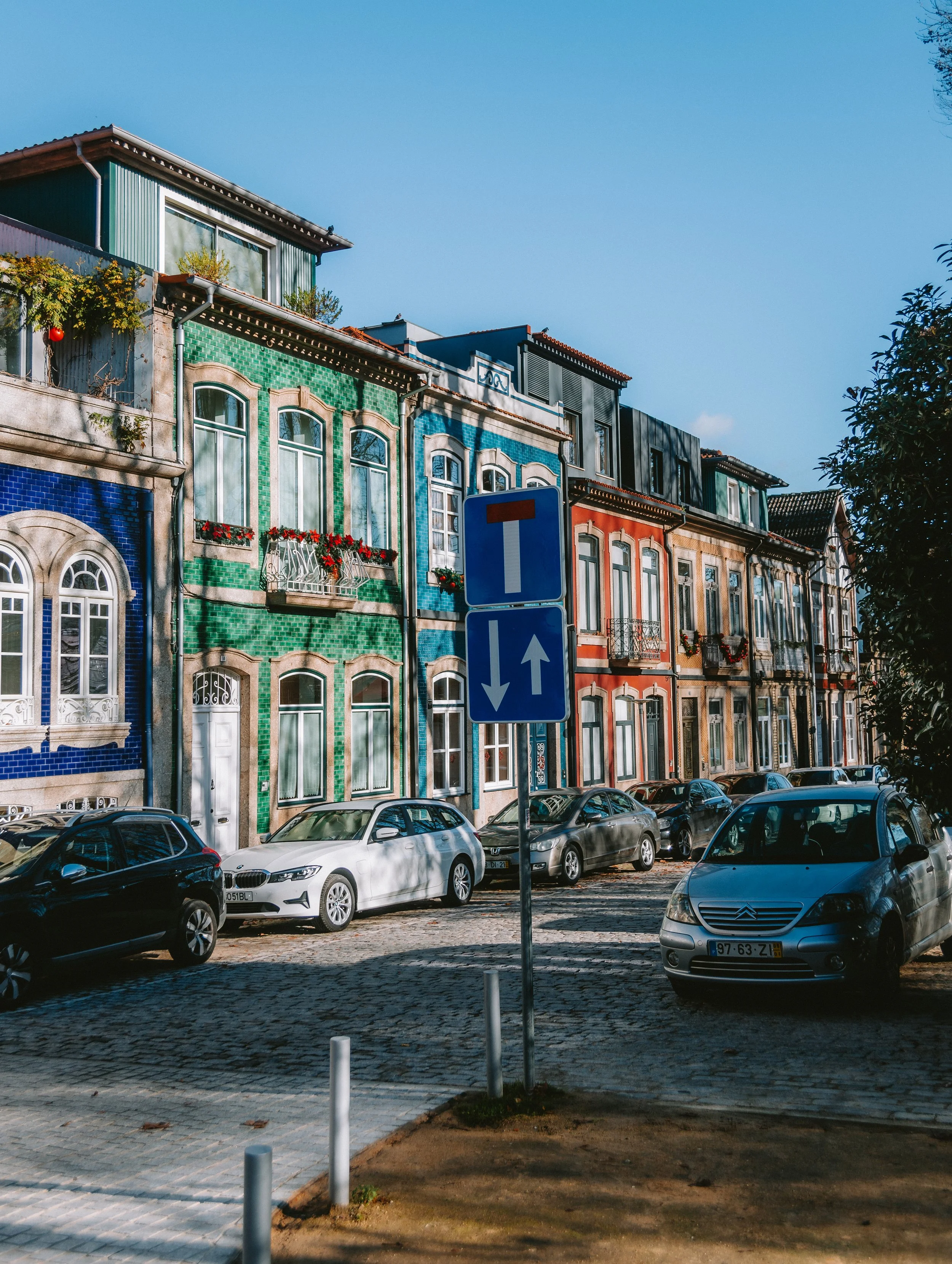 Colorful buildings with patterned facades and a row of parked cars along a cobblestone street, with a blue one-way traffic sign in the foreground under a clear blue sky.