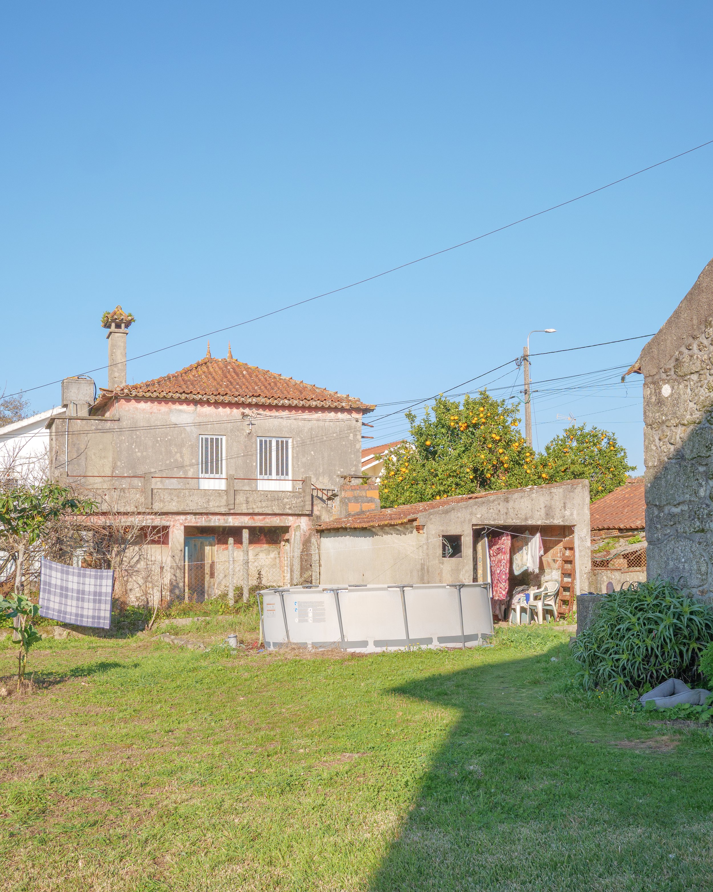 An outdoor backyard with a grassy lawn, a small shed with hanging laundry, a pool cover, and a two-story house with a tiled roof and a chimney.