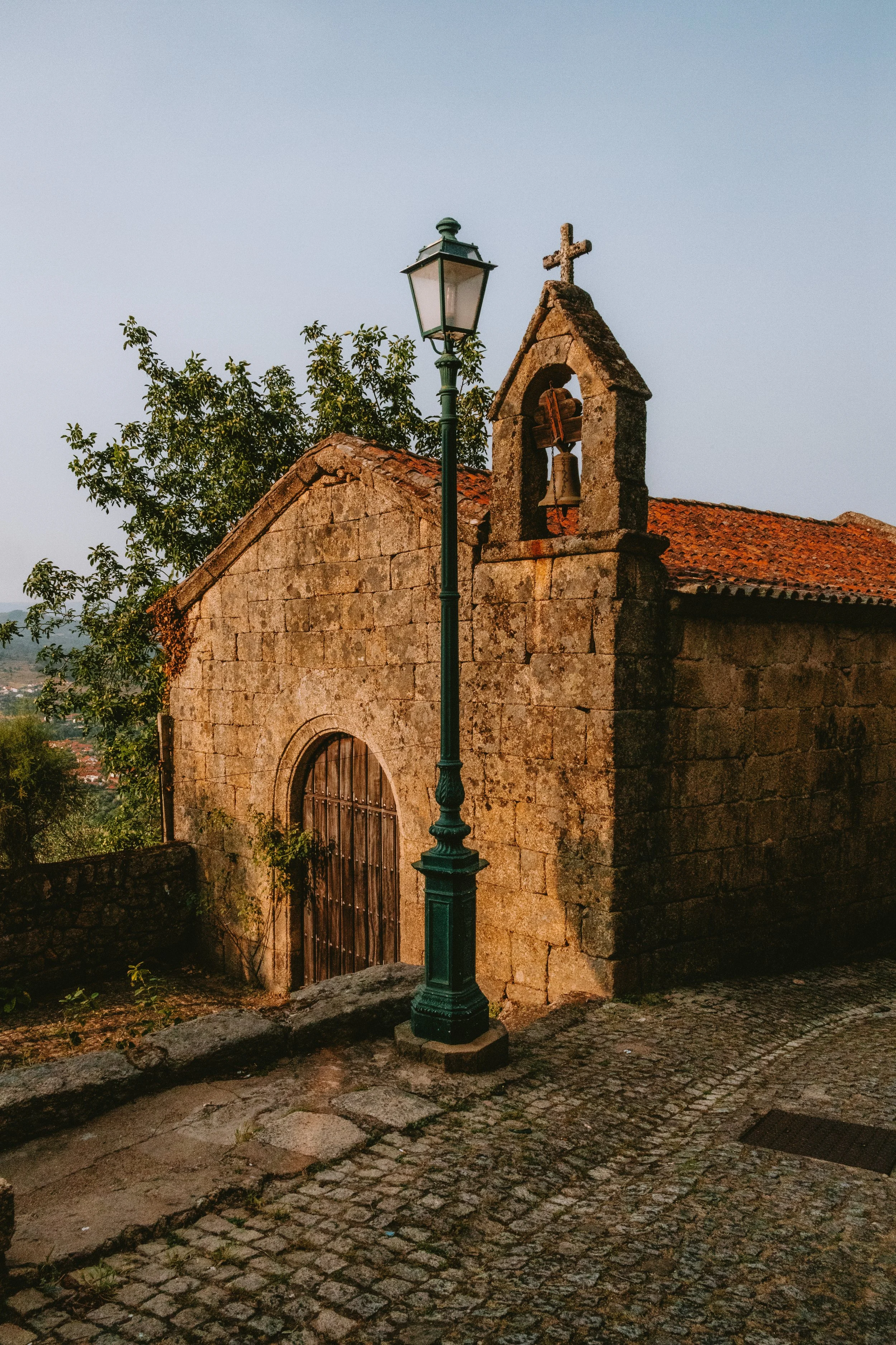 A small stone church with a wooden door, a bell tower with a cross on top, and a vintage street lamp in the foreground, illuminated by warm sunlight.