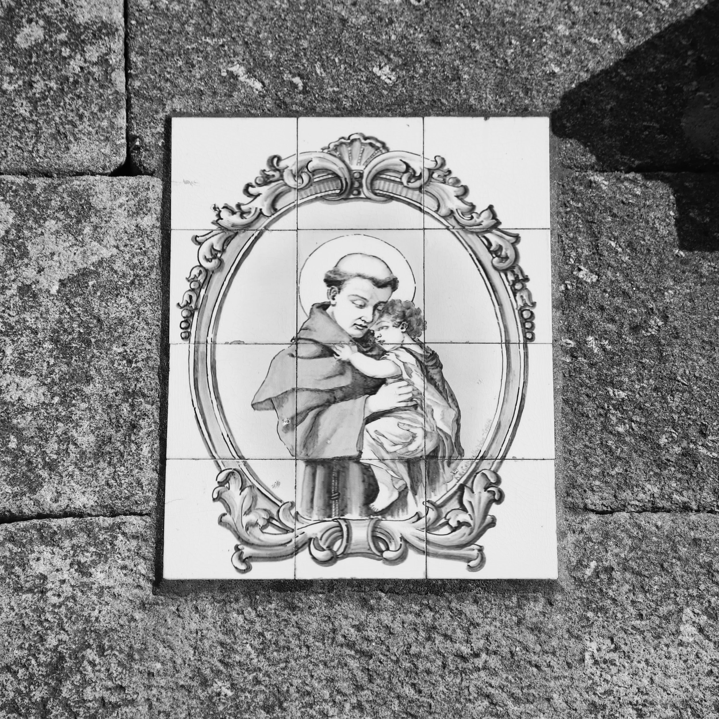 Black and white image of a ceramic tile with a religious depiction of a saint holding a child, surrounded by an ornate frame, laid on a stone surface.