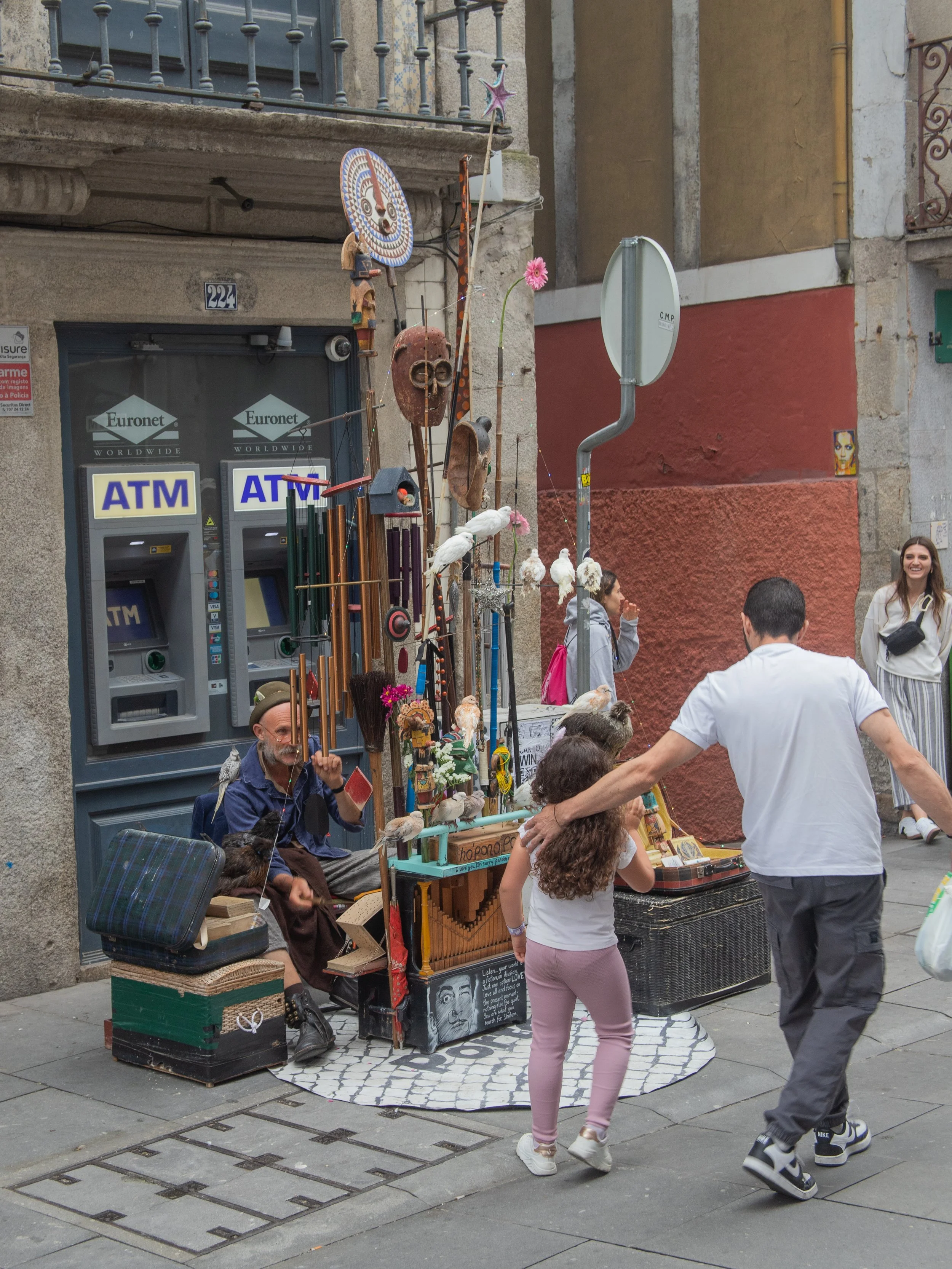 Street performer sitting next to a music box display with masks and figurines, while a man and girl interact nearby in an urban setting with people walking past.