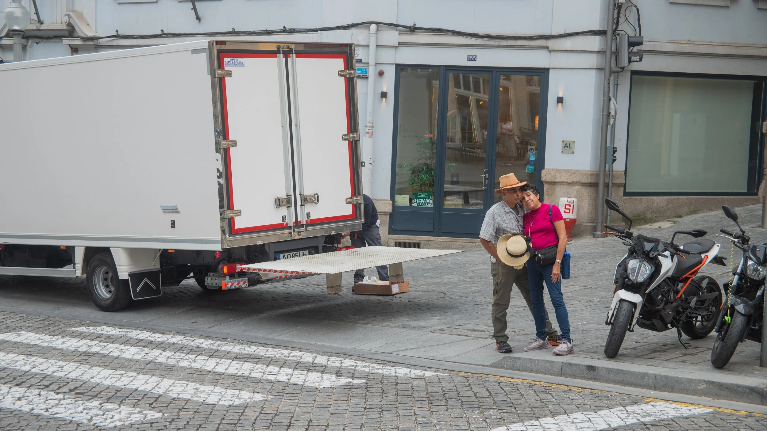 Two elderly people, a man and a woman, standing close together on a cobblestone street next to a motorcycle. The man is wearing a straw hat and casual clothes, holding a hat in his hand. The woman is dressed in a bright pink top and jeans, leaning he