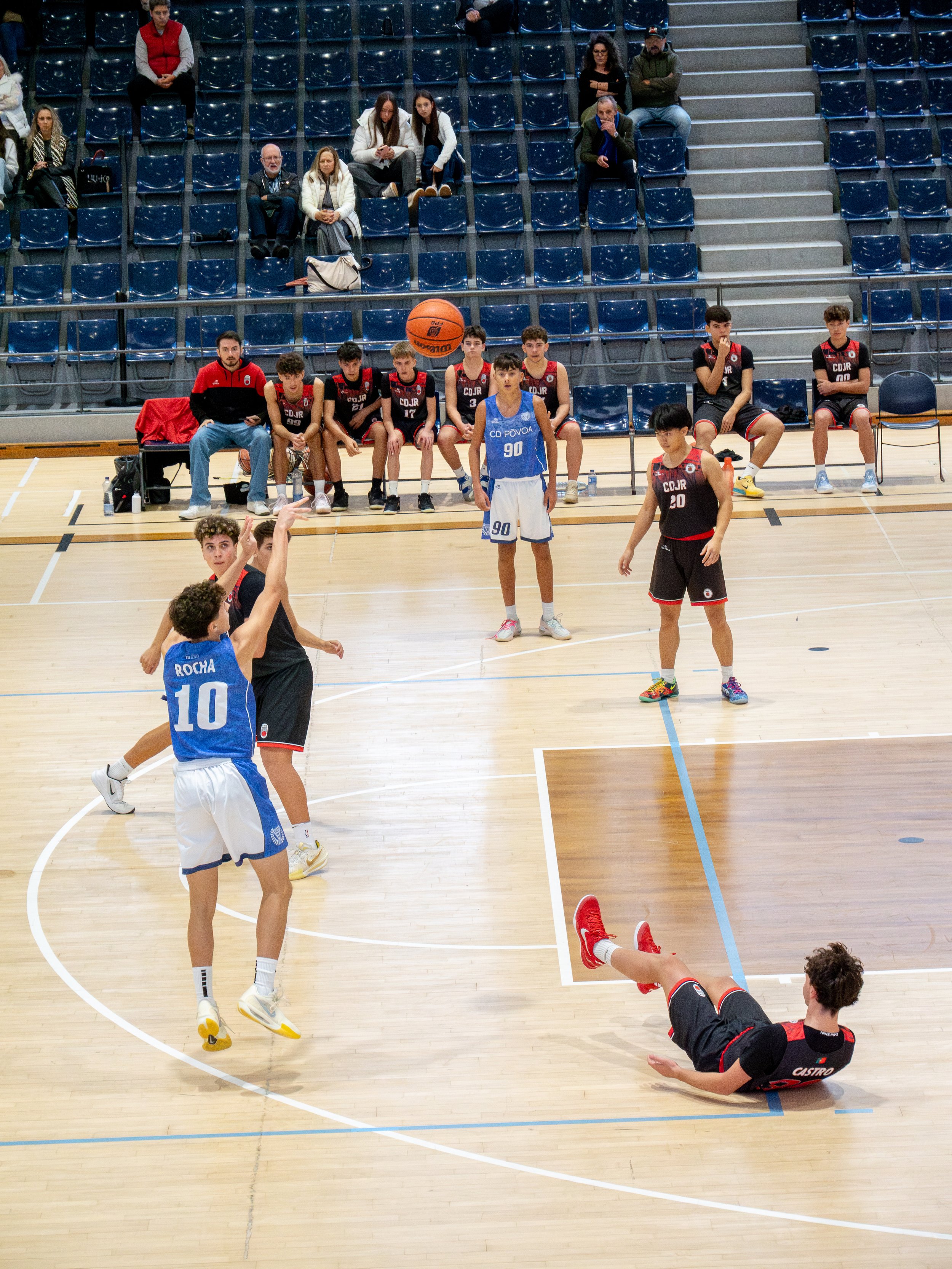 Young basketball players compete on an indoor court during a game, with some players shooting and others observing, while spectators watch from the bleachers.