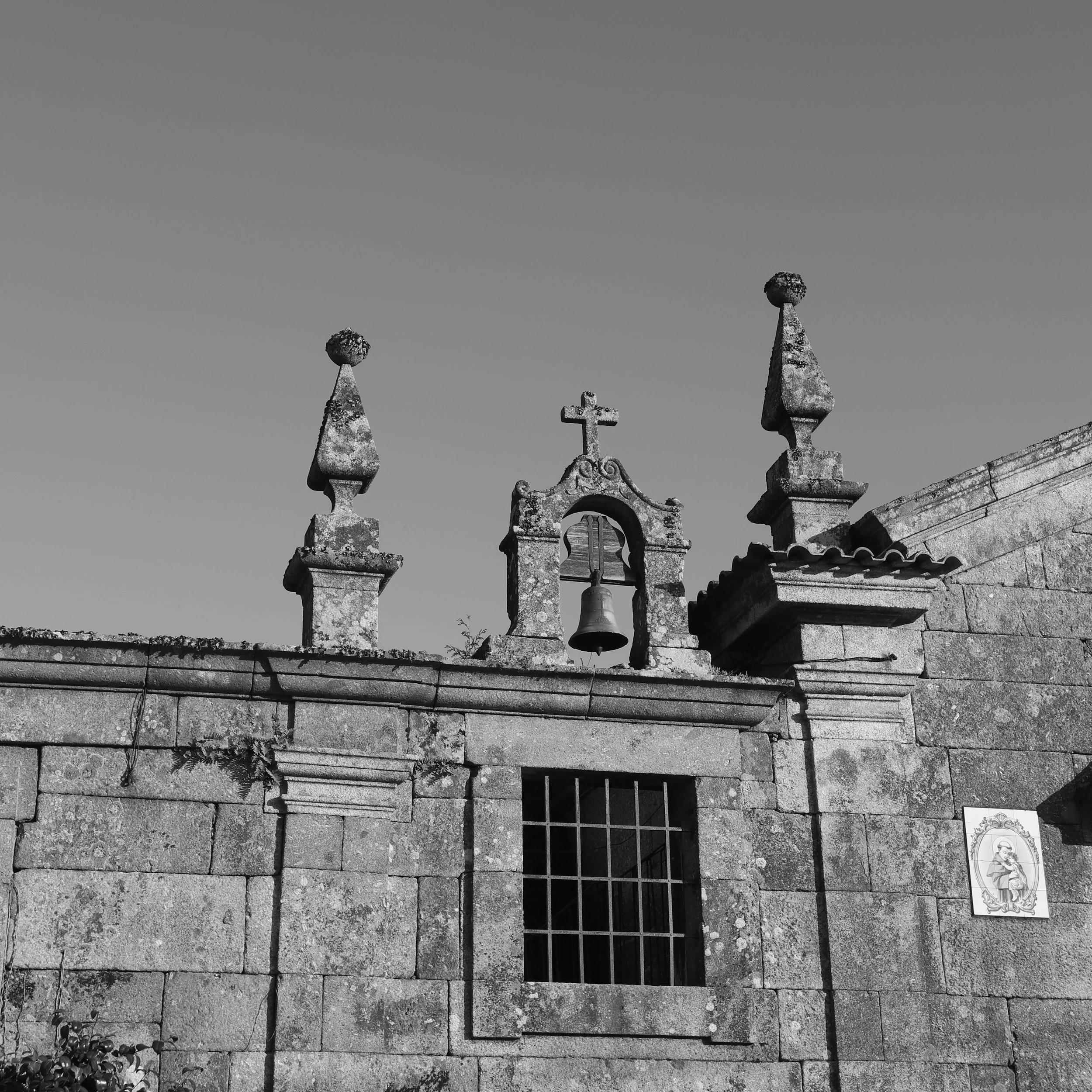 Black and white photo of a stone church wall with a bell in a niche, topped with a cross. Two decorative finials are on the roof. A small framed religious picture is on the right side of the wall.