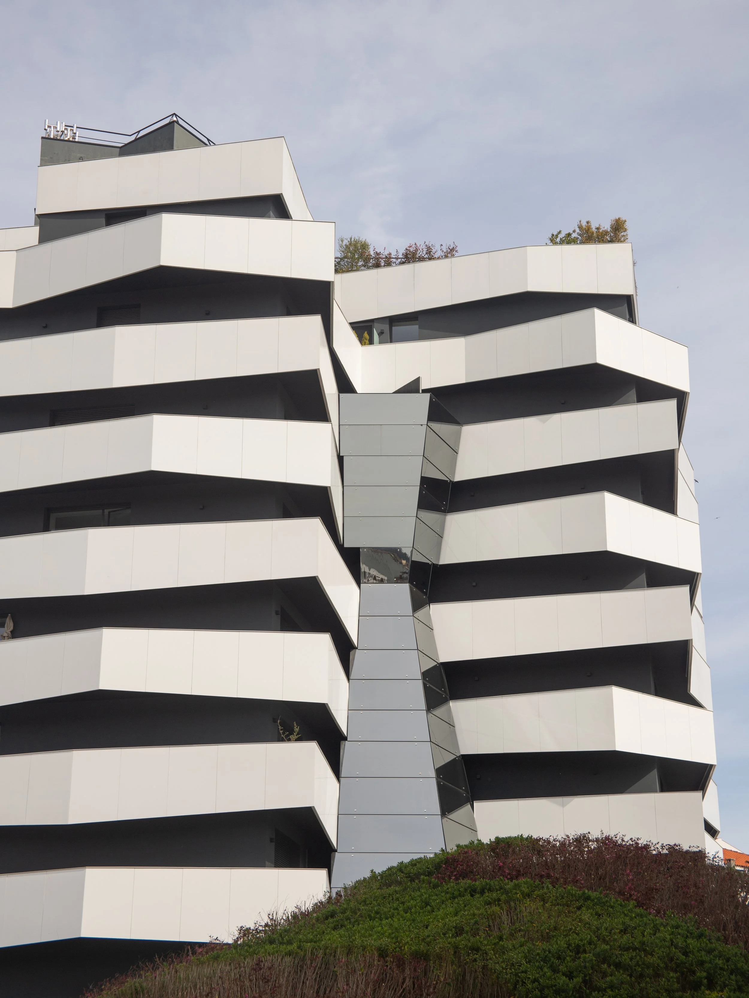 Modern multi-story building with staggered balcony design and a central glass elevator shaft, surrounded by greenery.