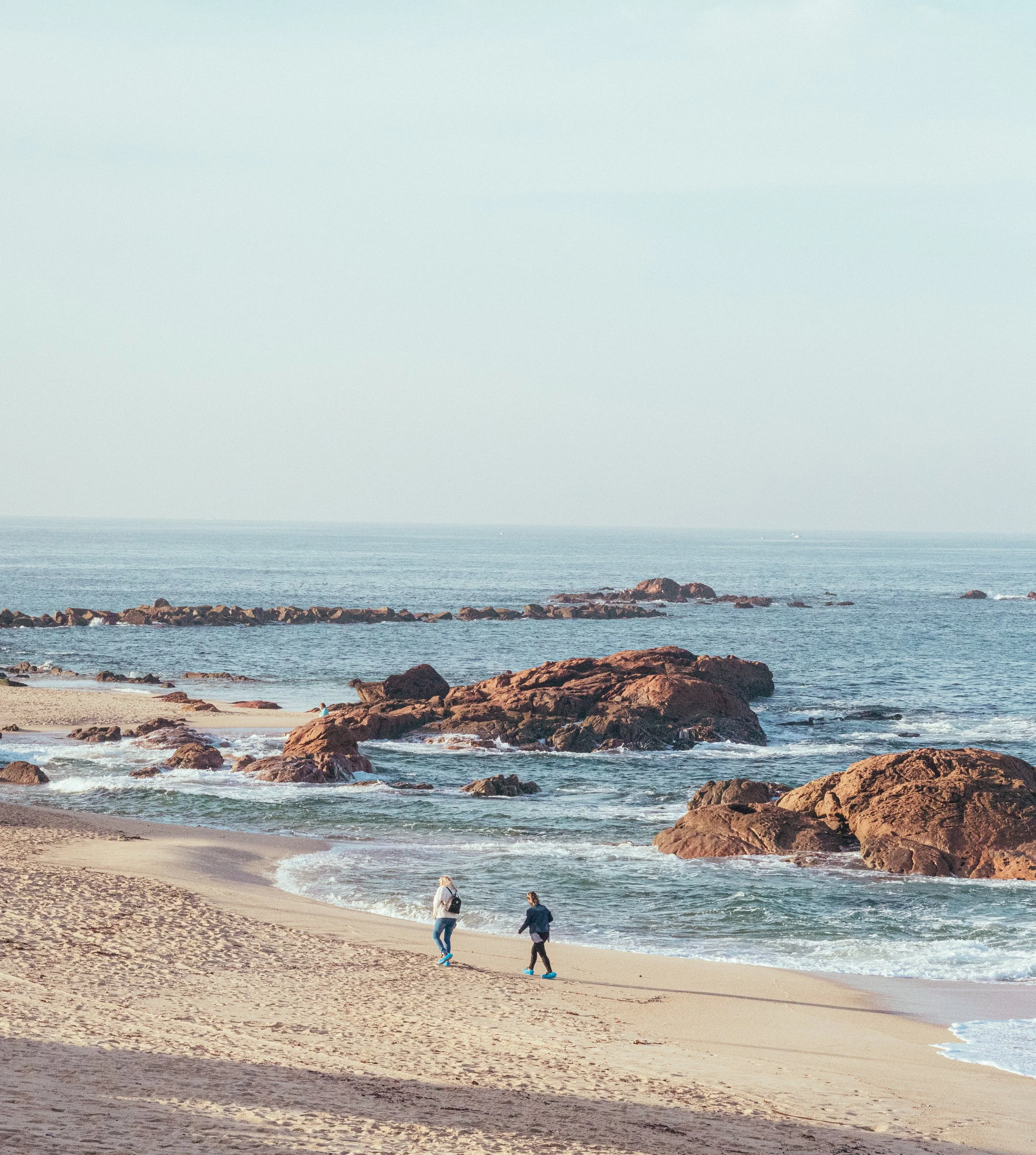 Two people walking along a sandy beach near large rocks and the ocean.