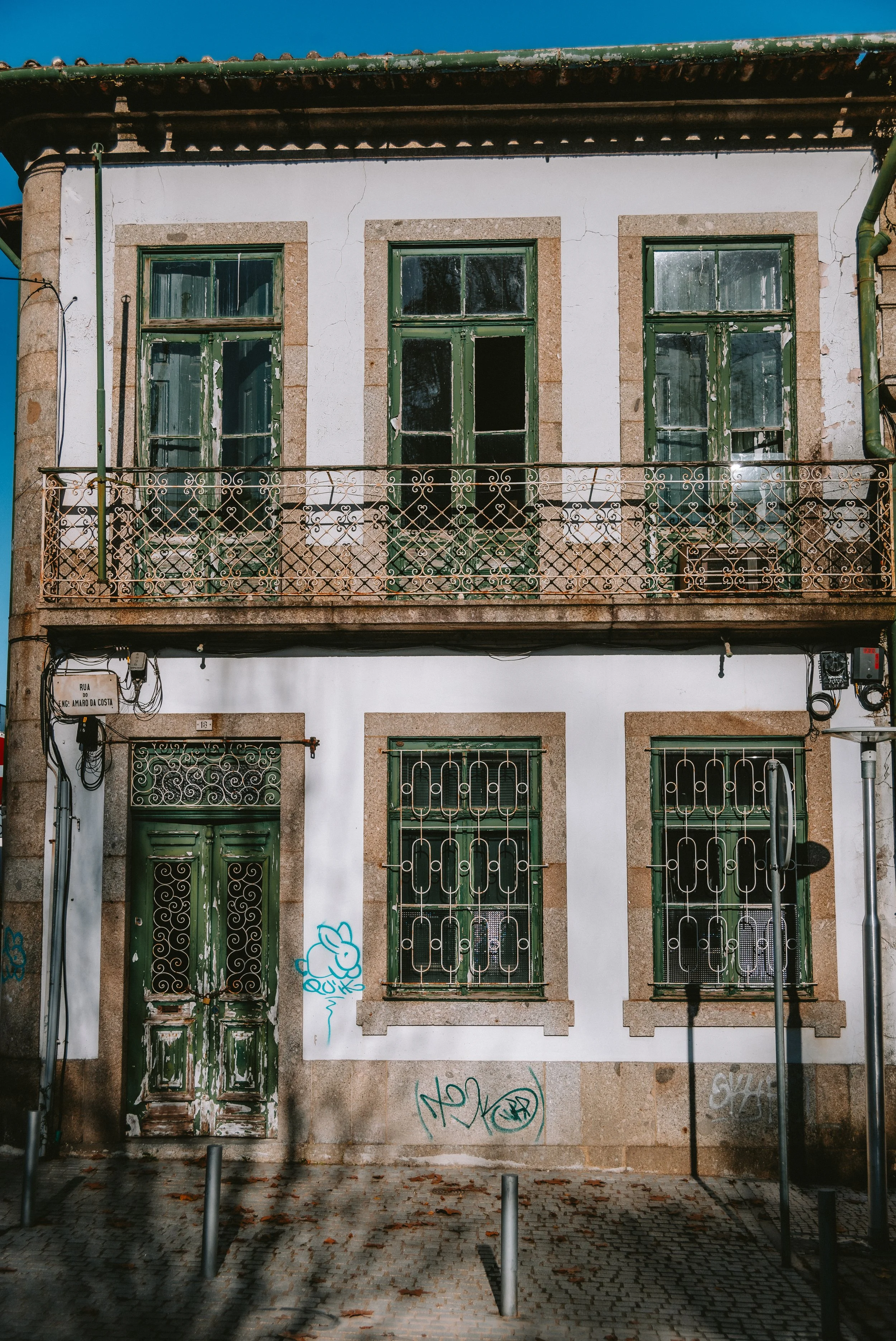 An old two-story building with white walls, green wooden doors, and windows with decorative metal bars. The building shows signs of wear, with peeling paint on the doors and windows, and some graffiti on the wall. There is a small balcony with a meta
