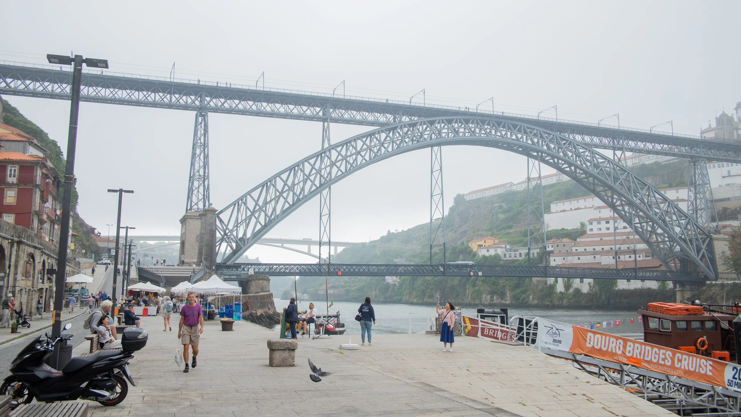 A scenic riverside view of the Douro River with the iconic Dom Luís I Bridge in Porto, Portugal. The area features people walking, sitting on benches, vendor stalls, and a boat dock. The bridge is a double-deck metal arch bridge spanning the river, w