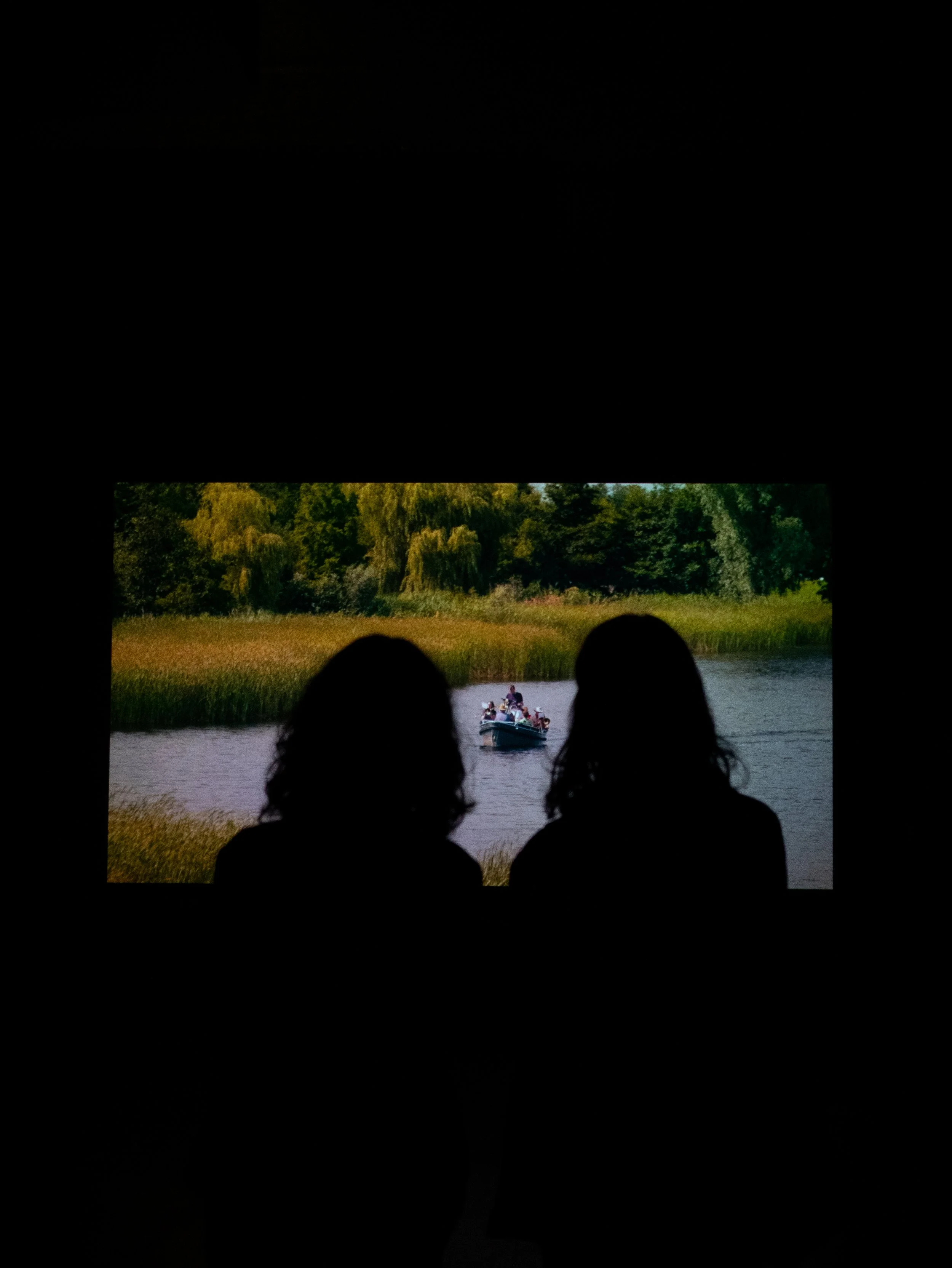 Silhouette of two women watching a movie on a screen showing a boat on a river with greenery in the background.