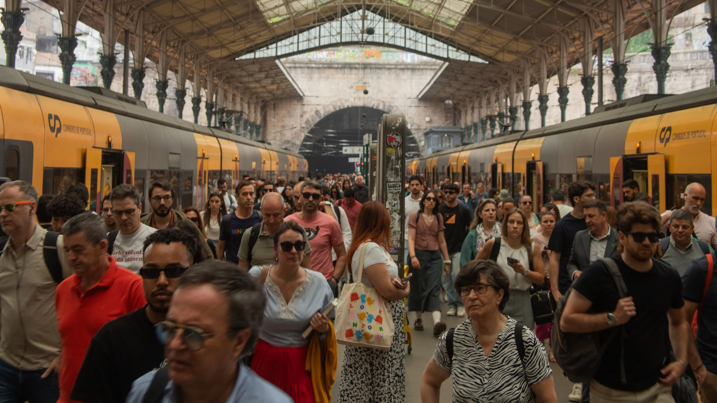 Crowded train station platform with people walking and waiting, yellow Portugal national rail trains on both sides, arched ceiling with metal beams, and a tunnel in the background.