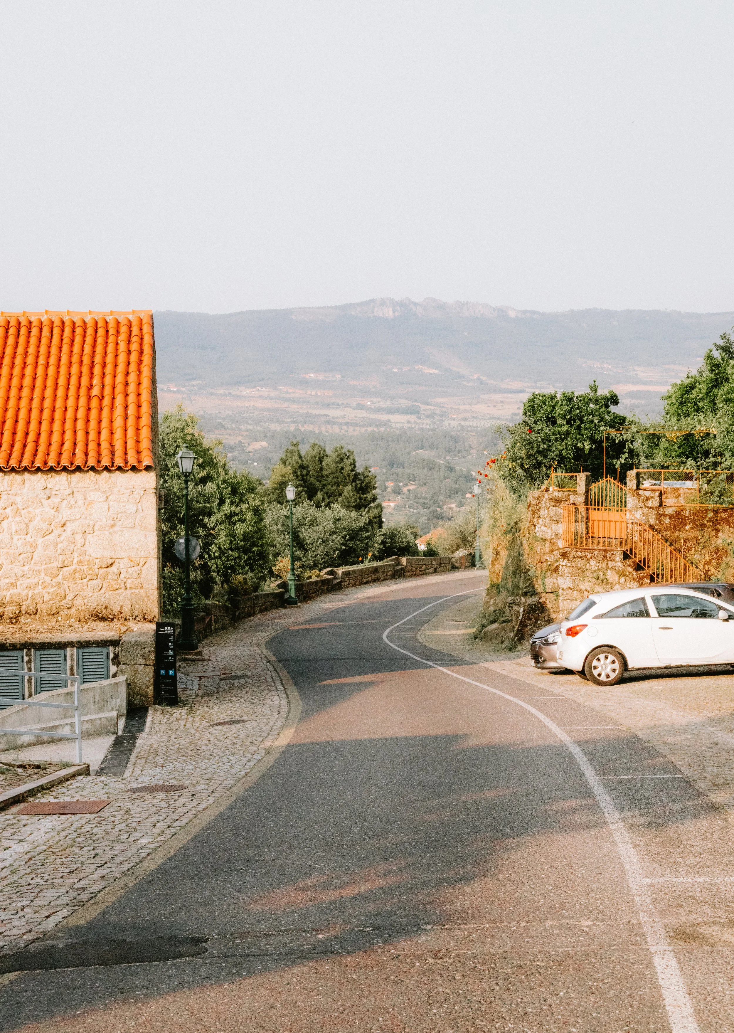 A winding road in a small town with a stone building with a red tile roof on the left, parked white car on the right, trees, street lamps, and a scenic view of mountains in the background.