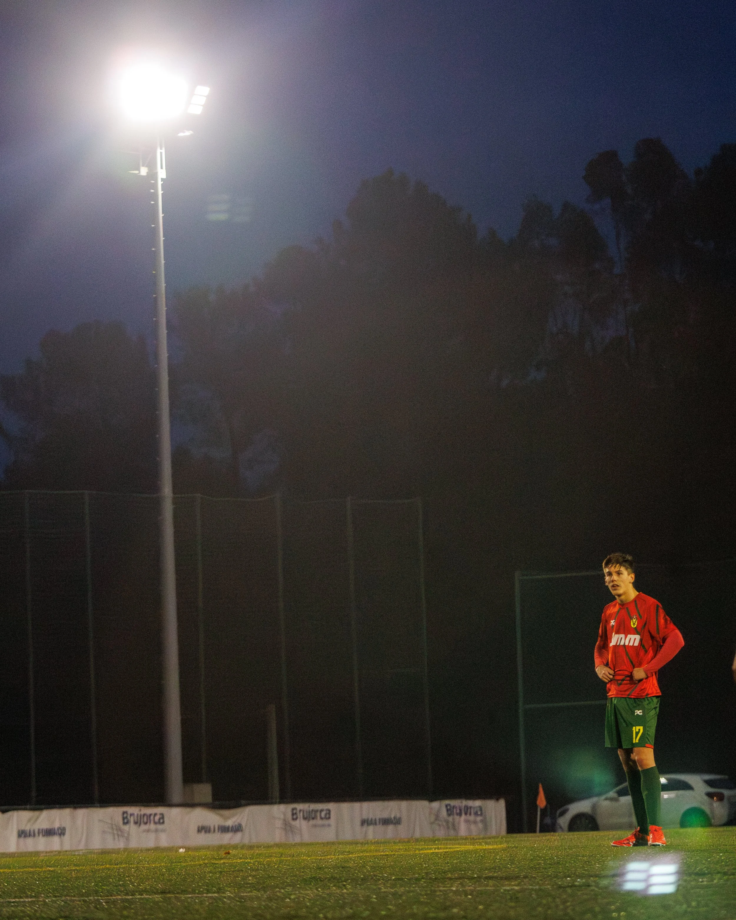 A soccer player standing on a field at night, illuminated by bright stadium lights, with trees and a fence in the background.