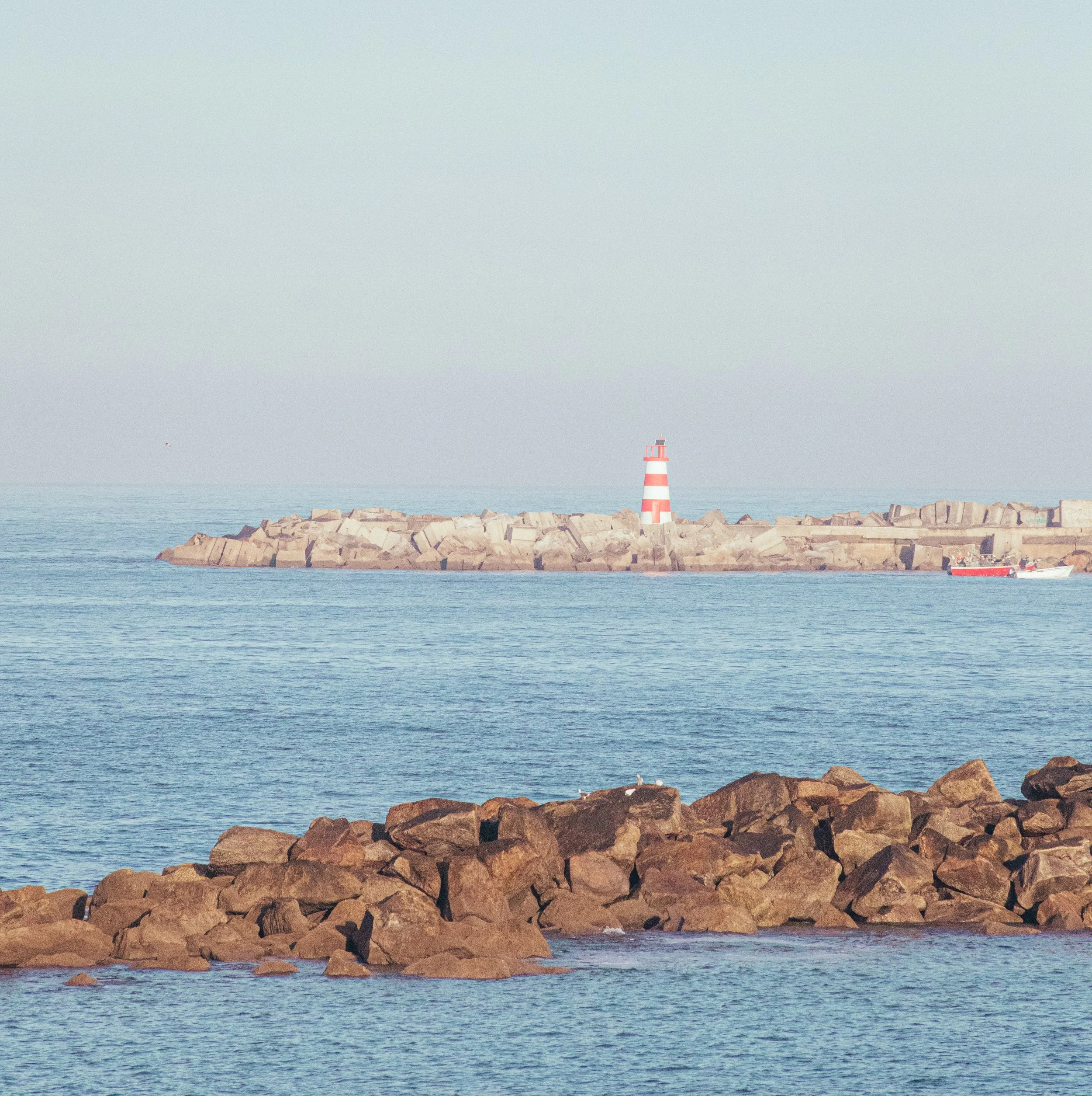 A lighthouse on a rocky breakwater in the ocean with a boat passing nearby. The scene is calm with a clear sky.