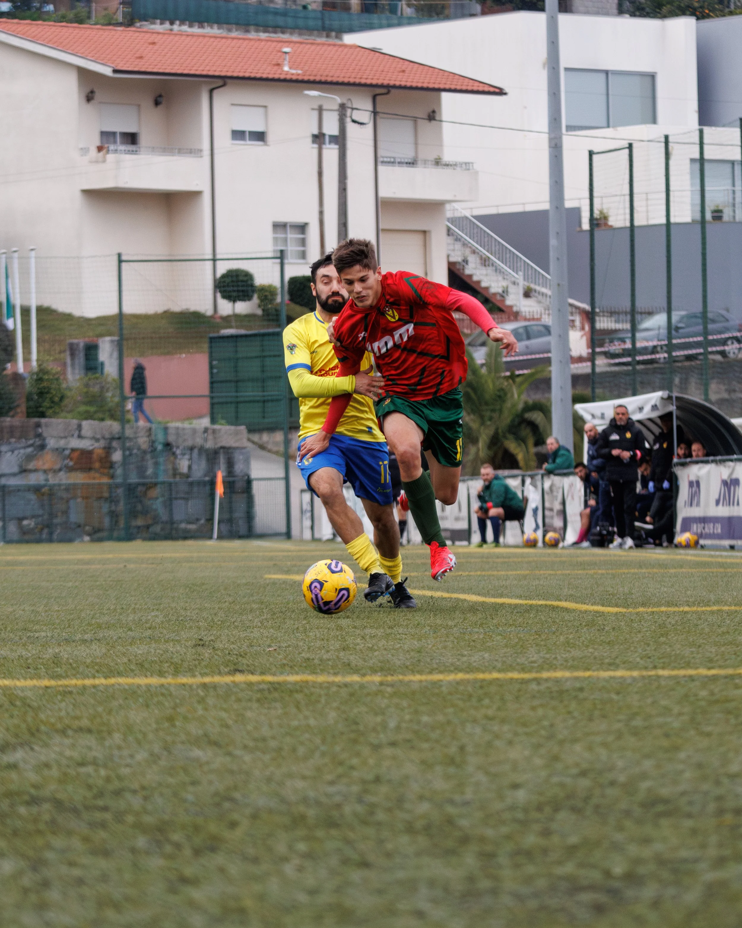 Two soccer players competing for the ball on a field, with spectators and a person sitting on the bench in the background.