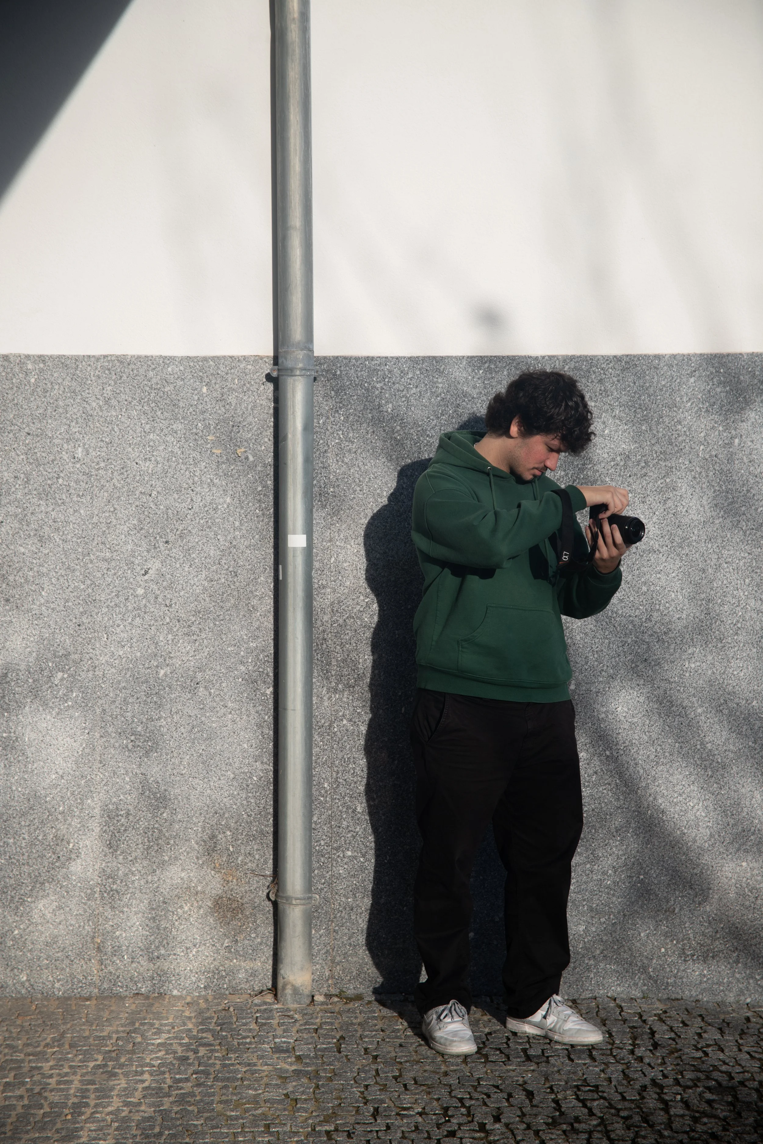 Young man standing against a wall, looking at a camera in his hand, with sunlight casting a shadow on the wall.