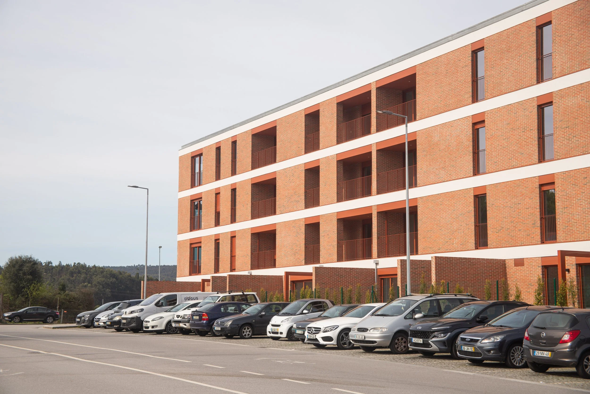 A modern brick apartment building with multiple balconies and a parking lot filled with various cars in front.