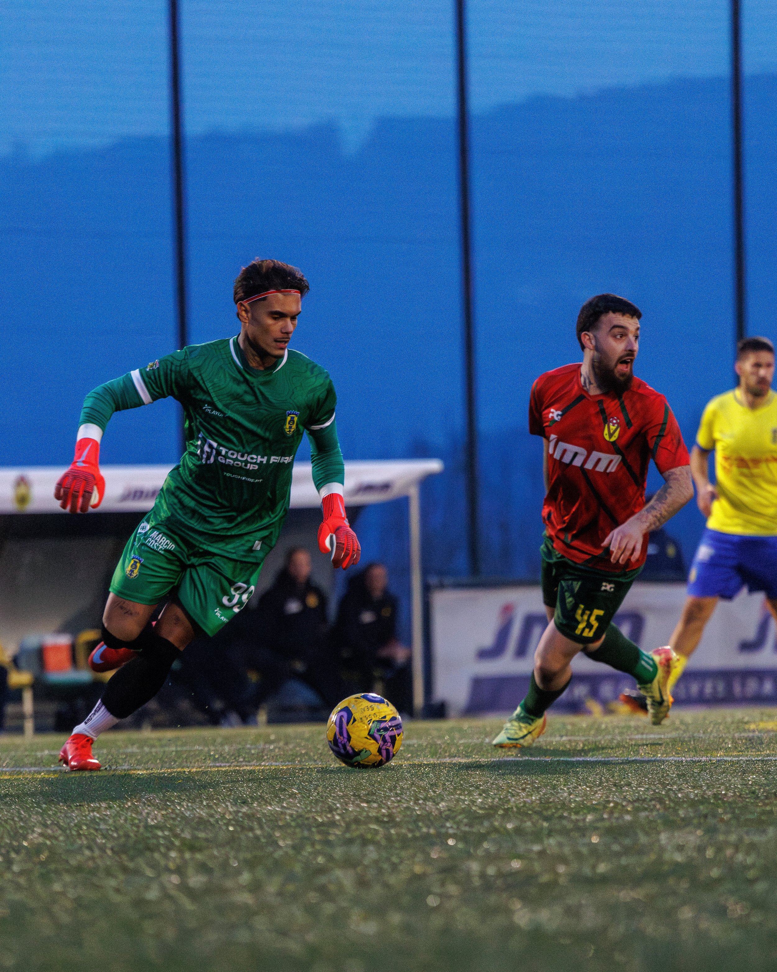 Soccer players chasing the ball during a match, with one player in a green kit and another in a red kit, on a grassy field with a blue background.
