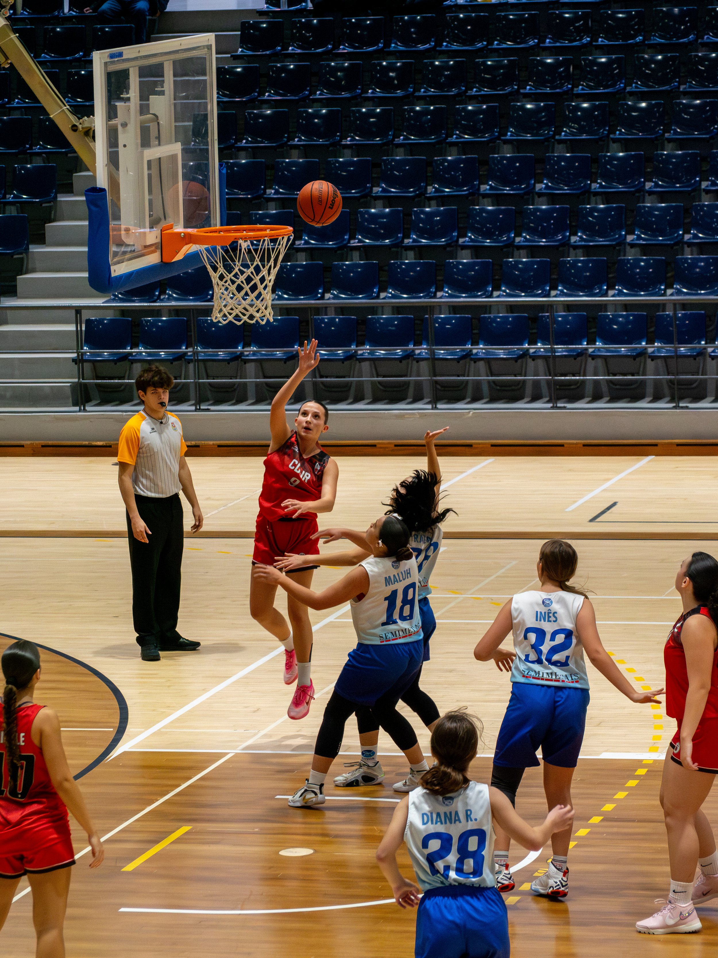Women playing basketball on an indoor court, with one player in red jumping for the ball near the hoop, while others in blue and white jerseys defend and observe.