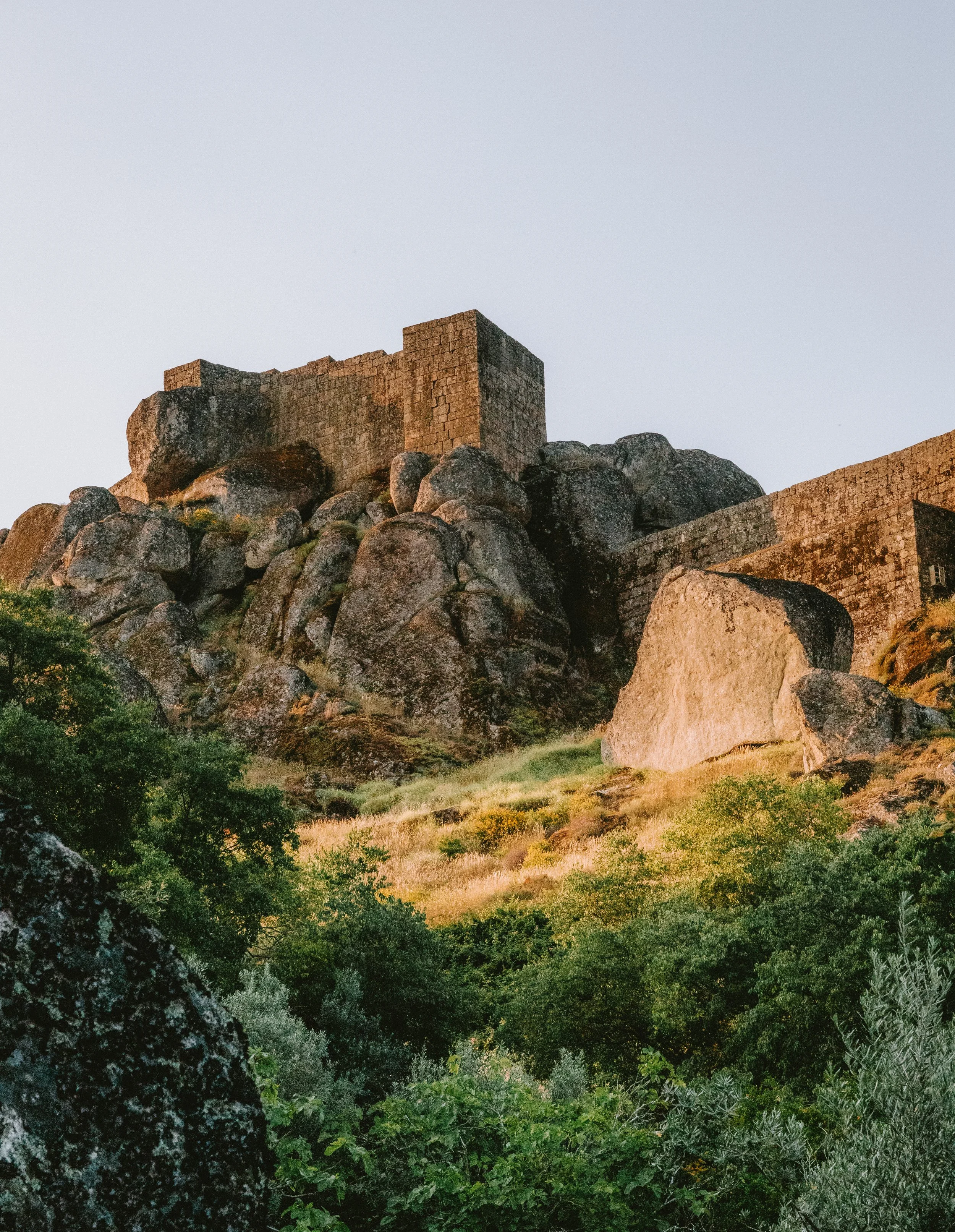 An ancient stone castle built on large boulders on a hilltop surrounded by lush green trees.
