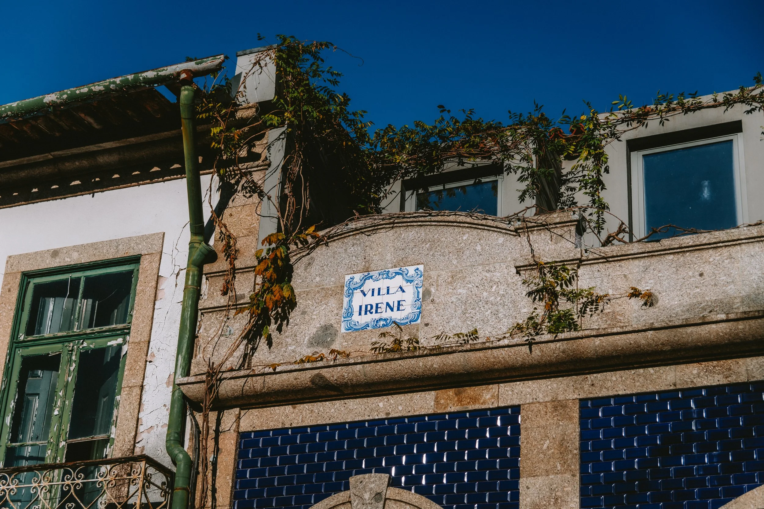Close-up of a building with a sign that reads 'Villa Irene,' featuring stone and blue tile facade, windows, and overgrown vines under clear blue sky.