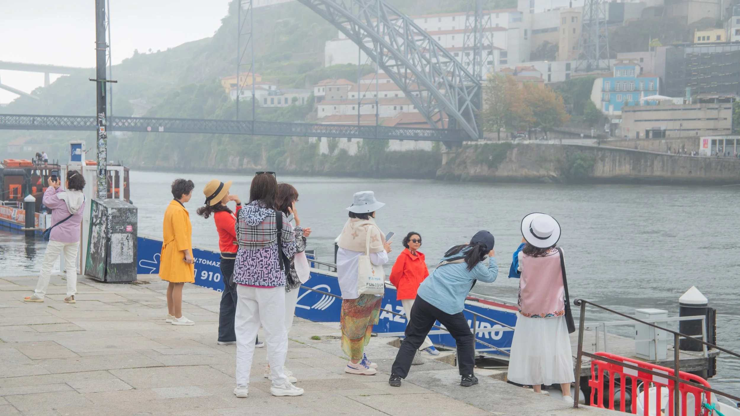 People standing on a riverside dock, with some looking at the water and one taking photos. A bridge and hillside with houses are visible in the background.