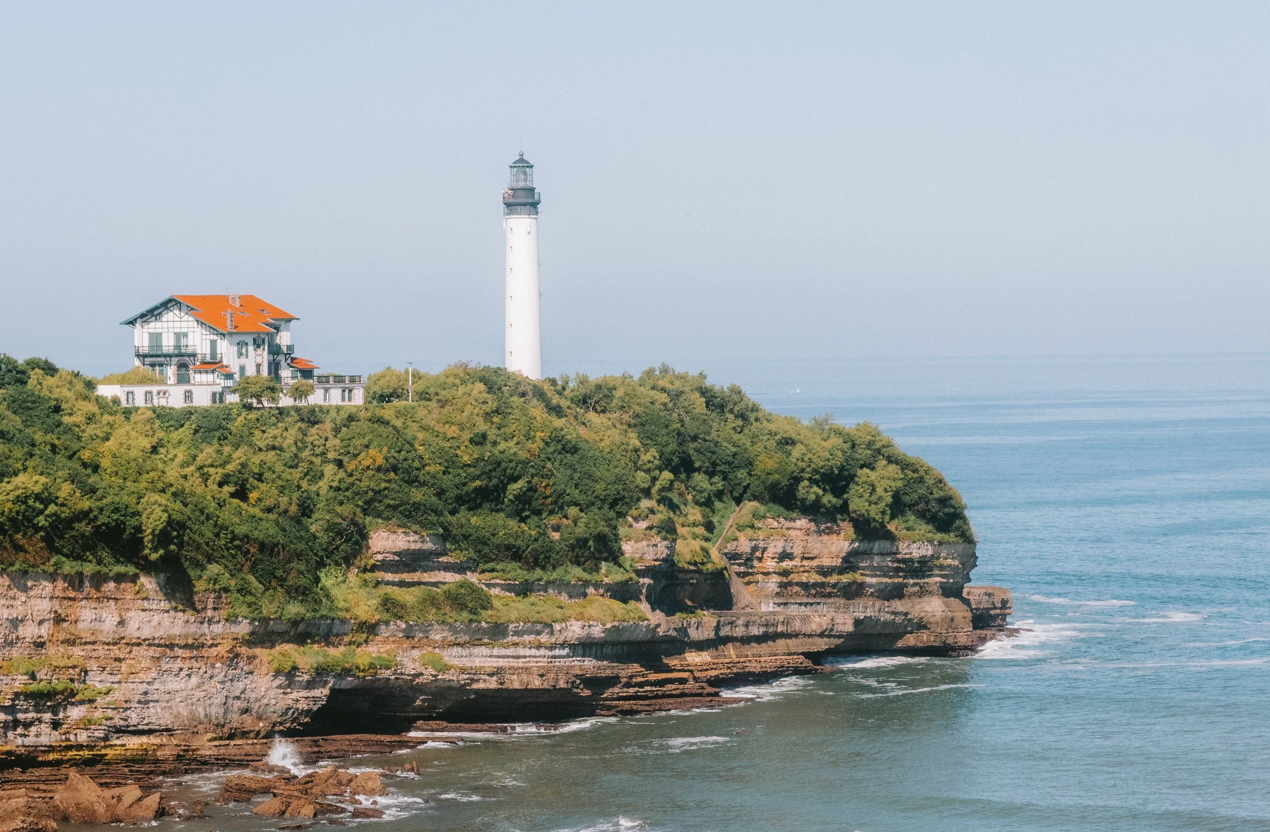 A lighthouse on a green, rocky cliff overlooking the ocean with a white house nearby, under a clear sky.