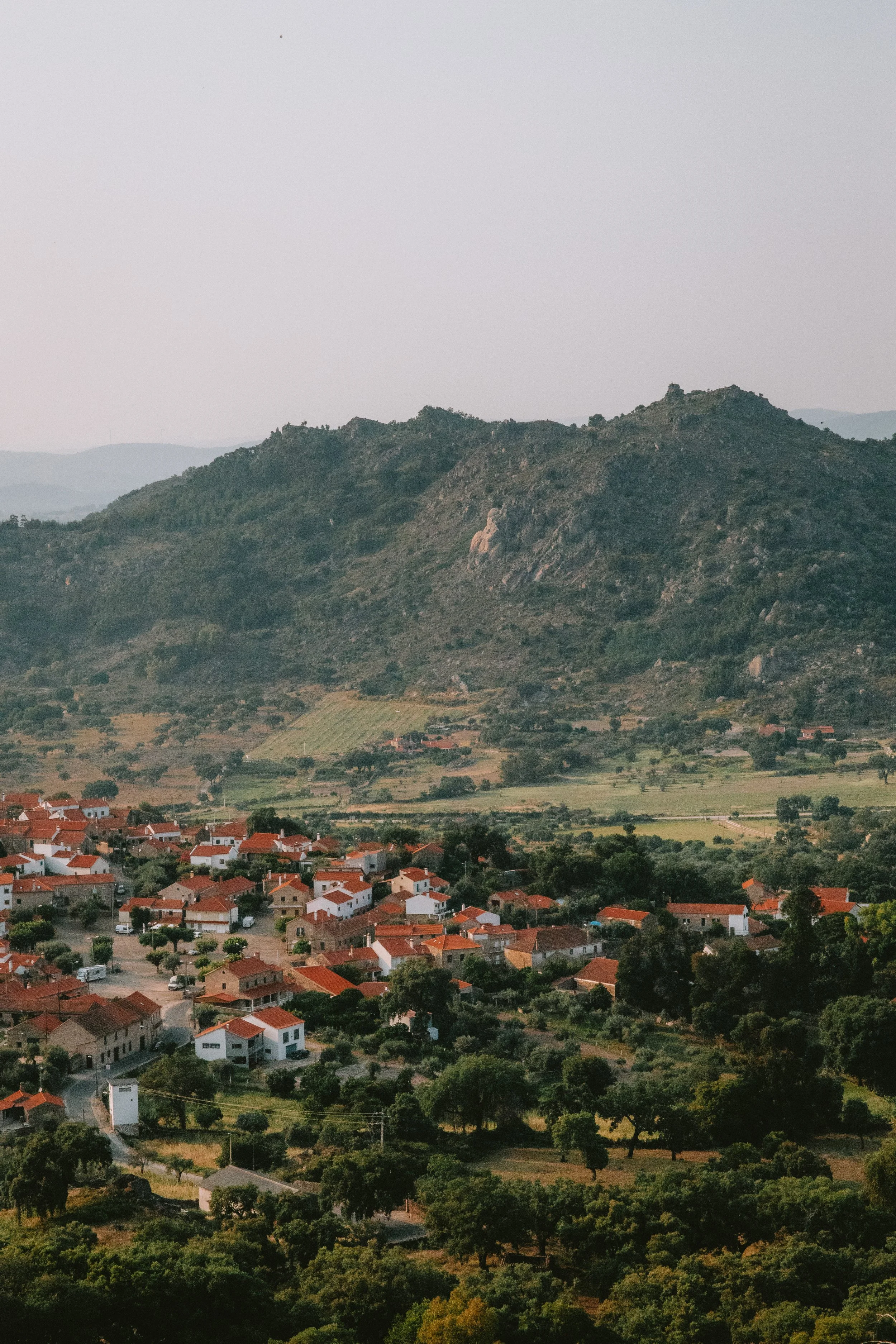 A scenic view of a small town with red-roofed houses and greenery, set against a backdrop of rolling hills and mountains.