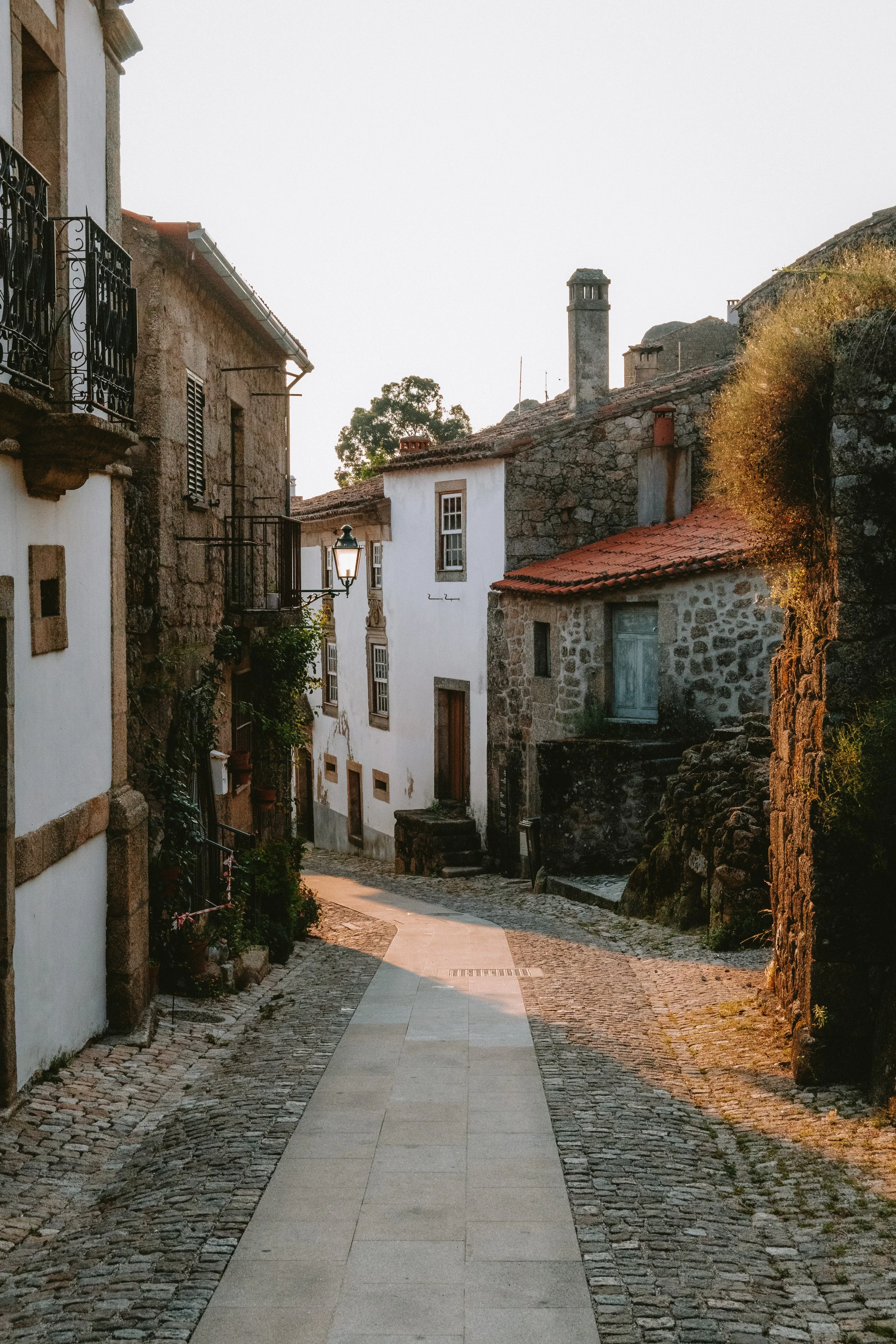 A narrow cobblestone street in an old European village, with stone and stucco buildings, some with wrought iron balconies, and a street lamp, bathed in warm sunlight.