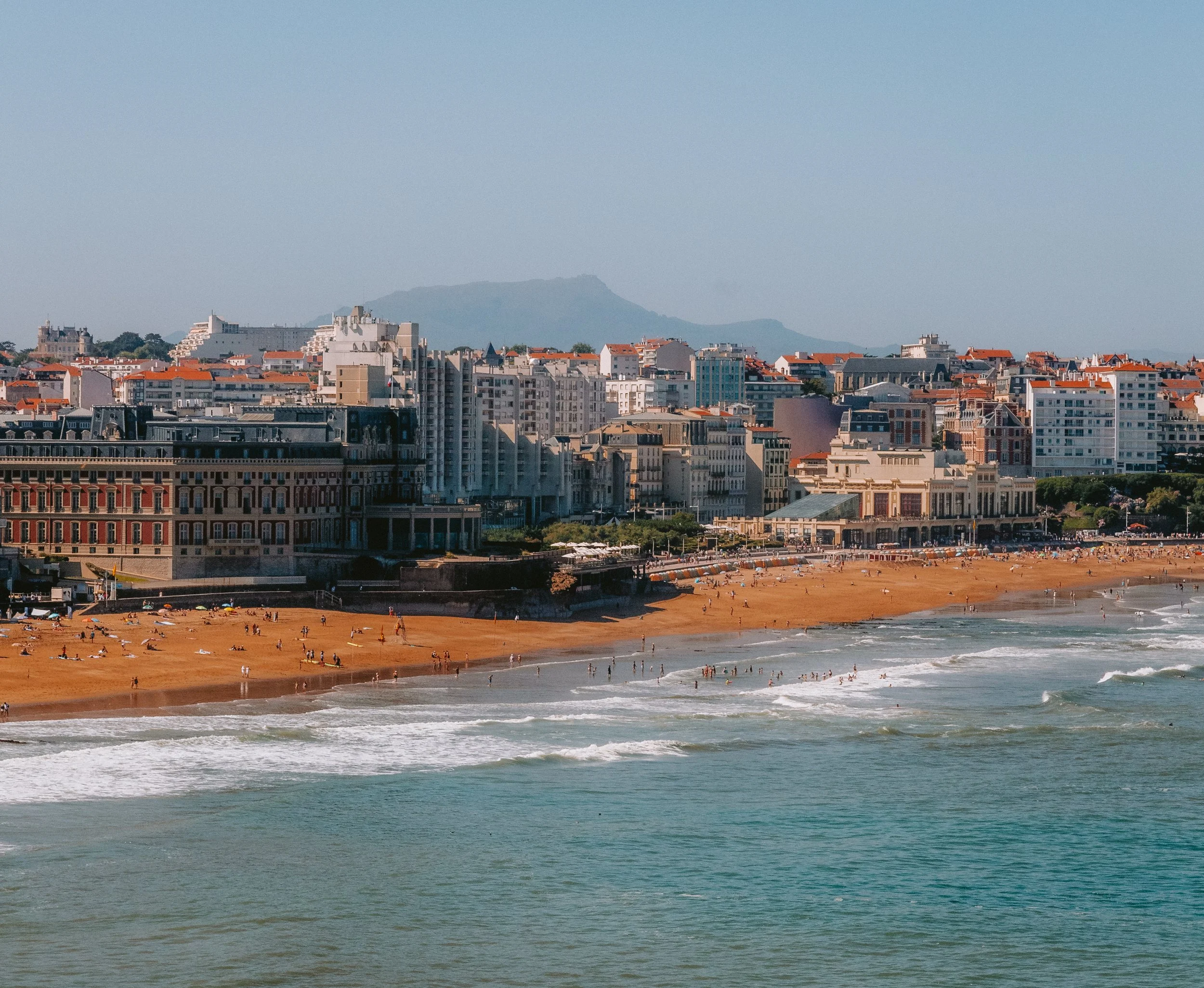 A city skyline with a beach in the foreground, people swimming and sunbathing, high-rise buildings, and a mountain in the background.