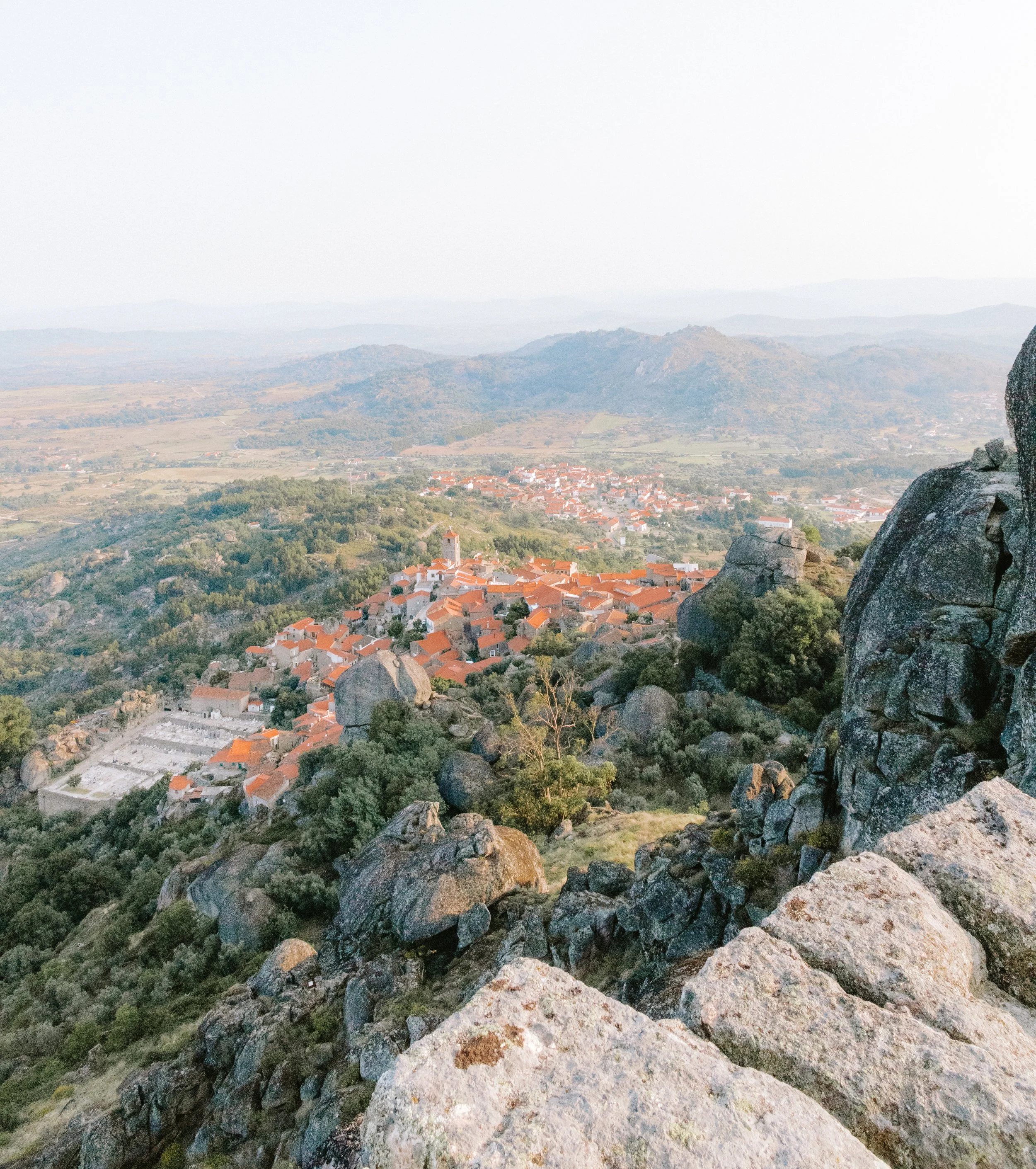 A scenic view of a small town with orange rooftops surrounded by rocky hills and distant mountains.