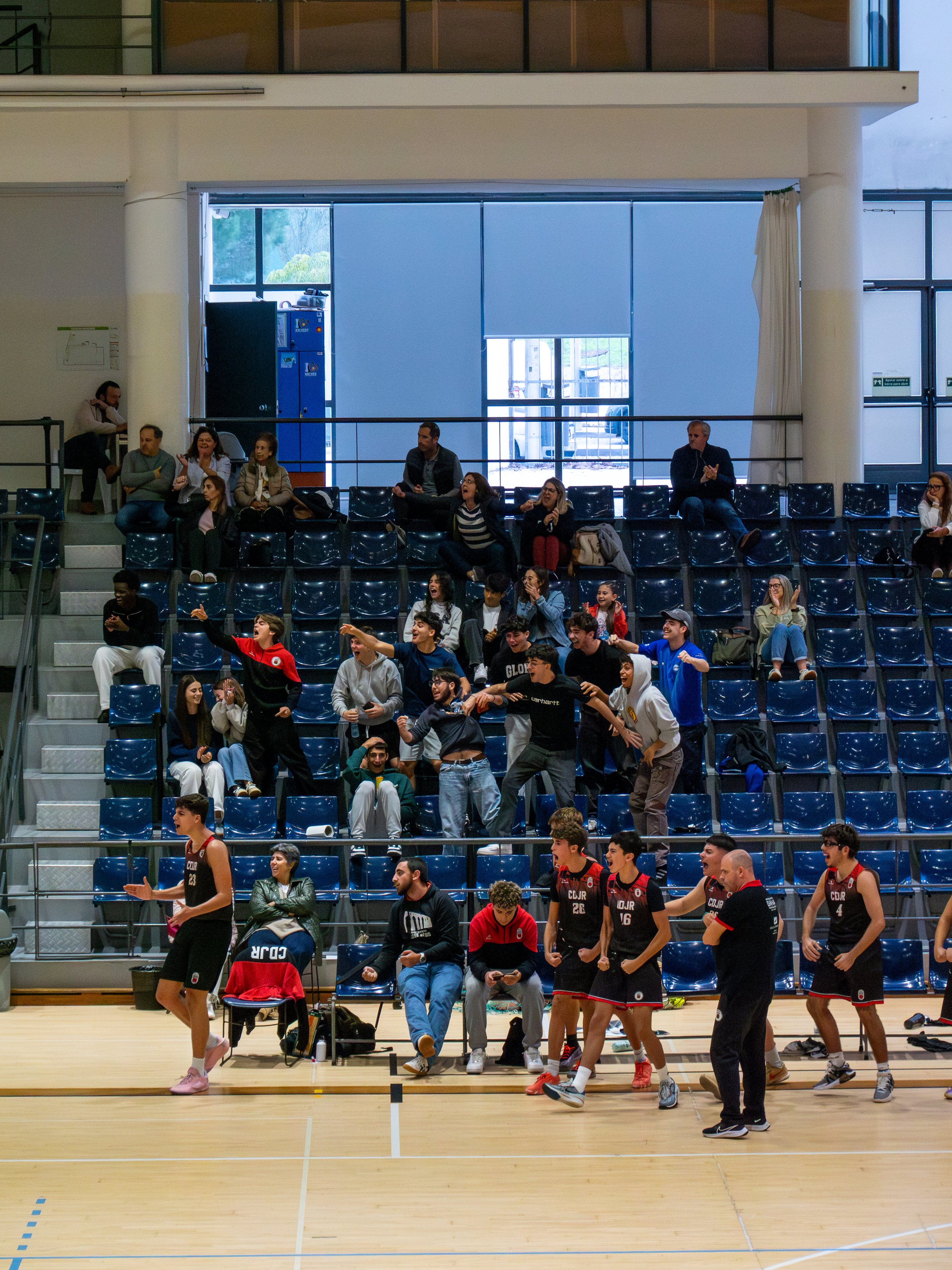 A basketball game in progress on an indoor court, with players in black uniforms and spectators in the stands, some standing and others sitting.