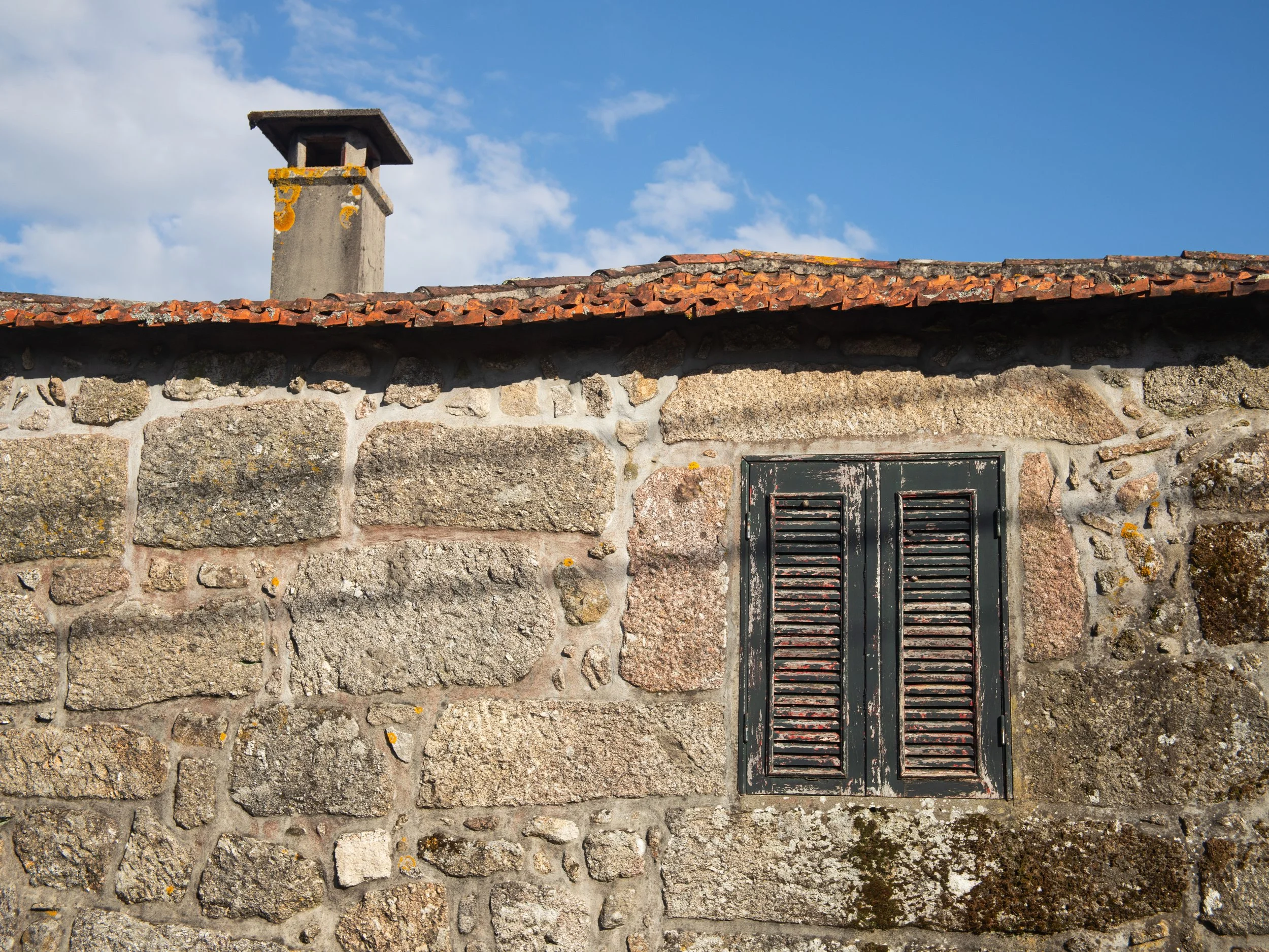 Stone wall with black window shutters and a tiled roof, chimney in the background, blue sky with clouds.