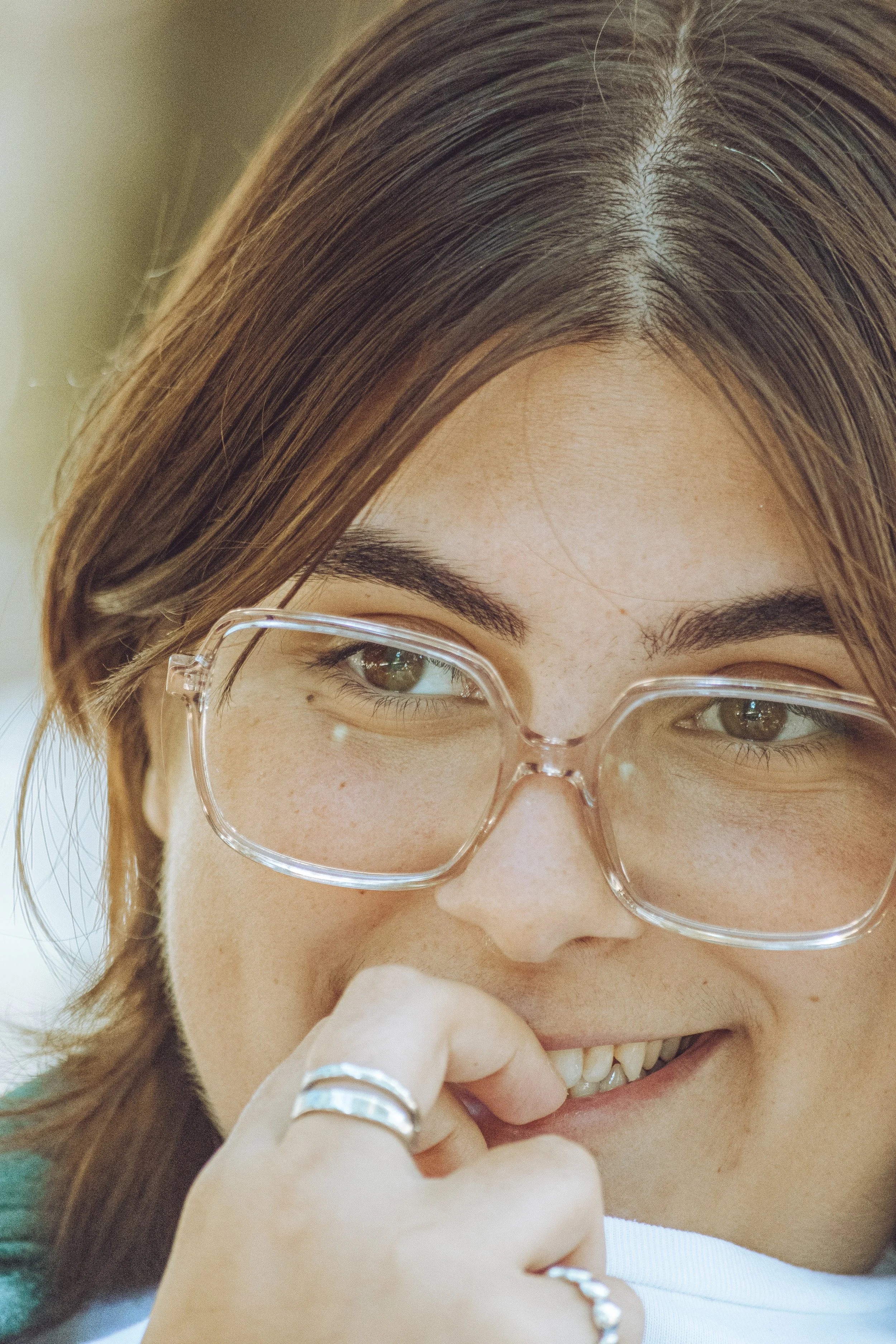 Close-up of a young woman with brown hair, wearing clear glasses, smiling softly while touching her chin, with rings on her fingers.