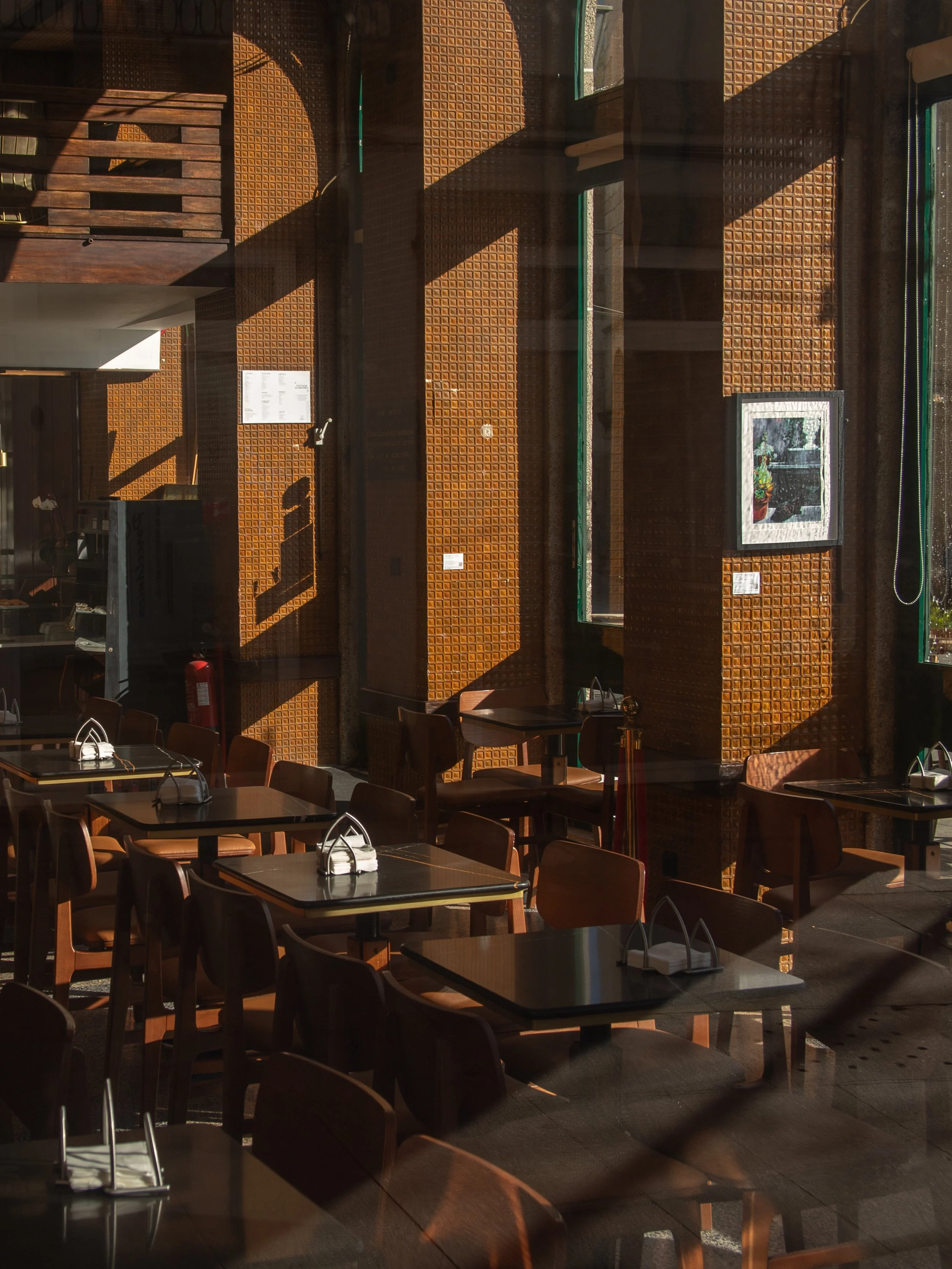 Empty restaurant with wooden tables and chairs, sunlight casting shadows through windows.