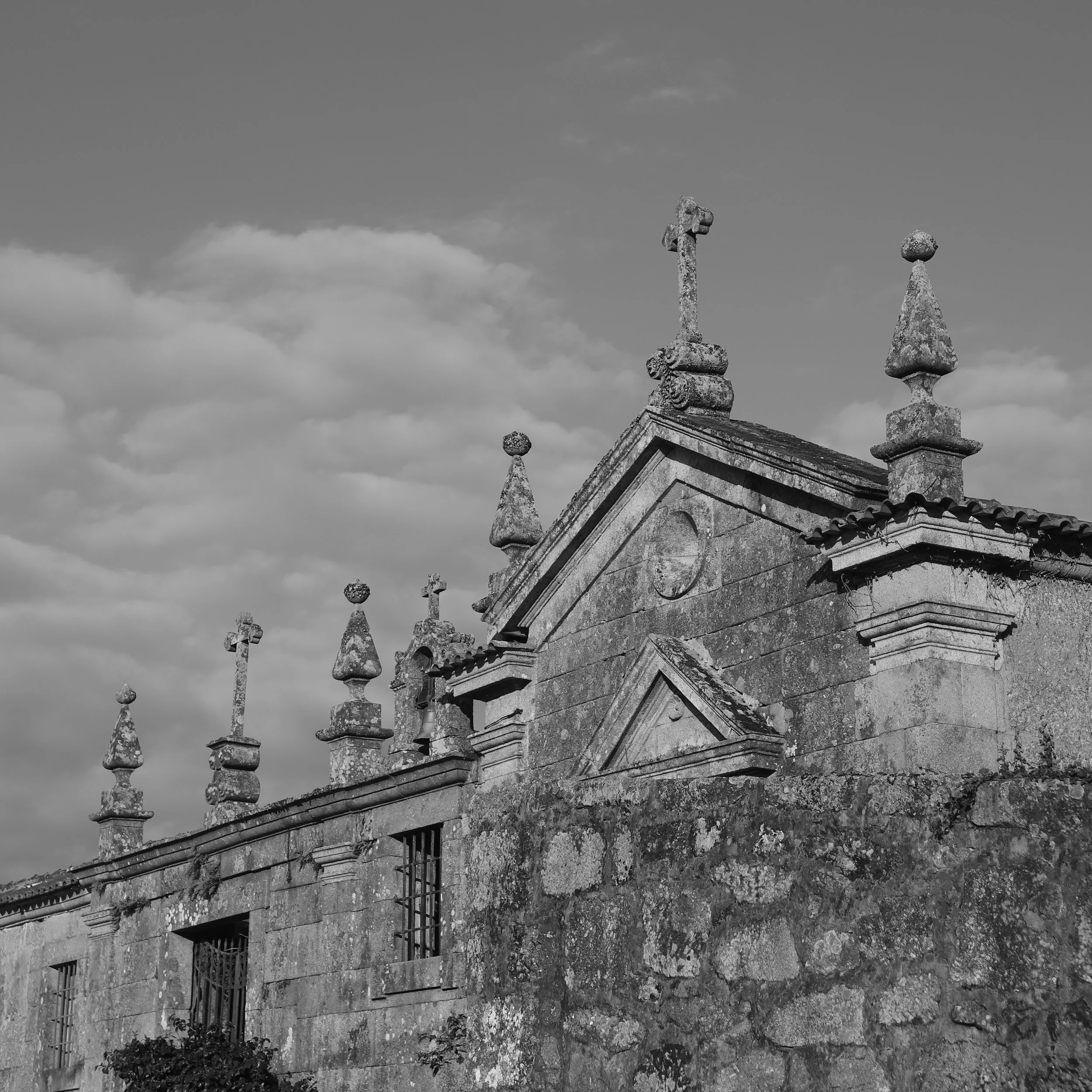 Black and white photo of an old stone church with decorative finials on the roof, some with crosses on top, under a cloudy sky.