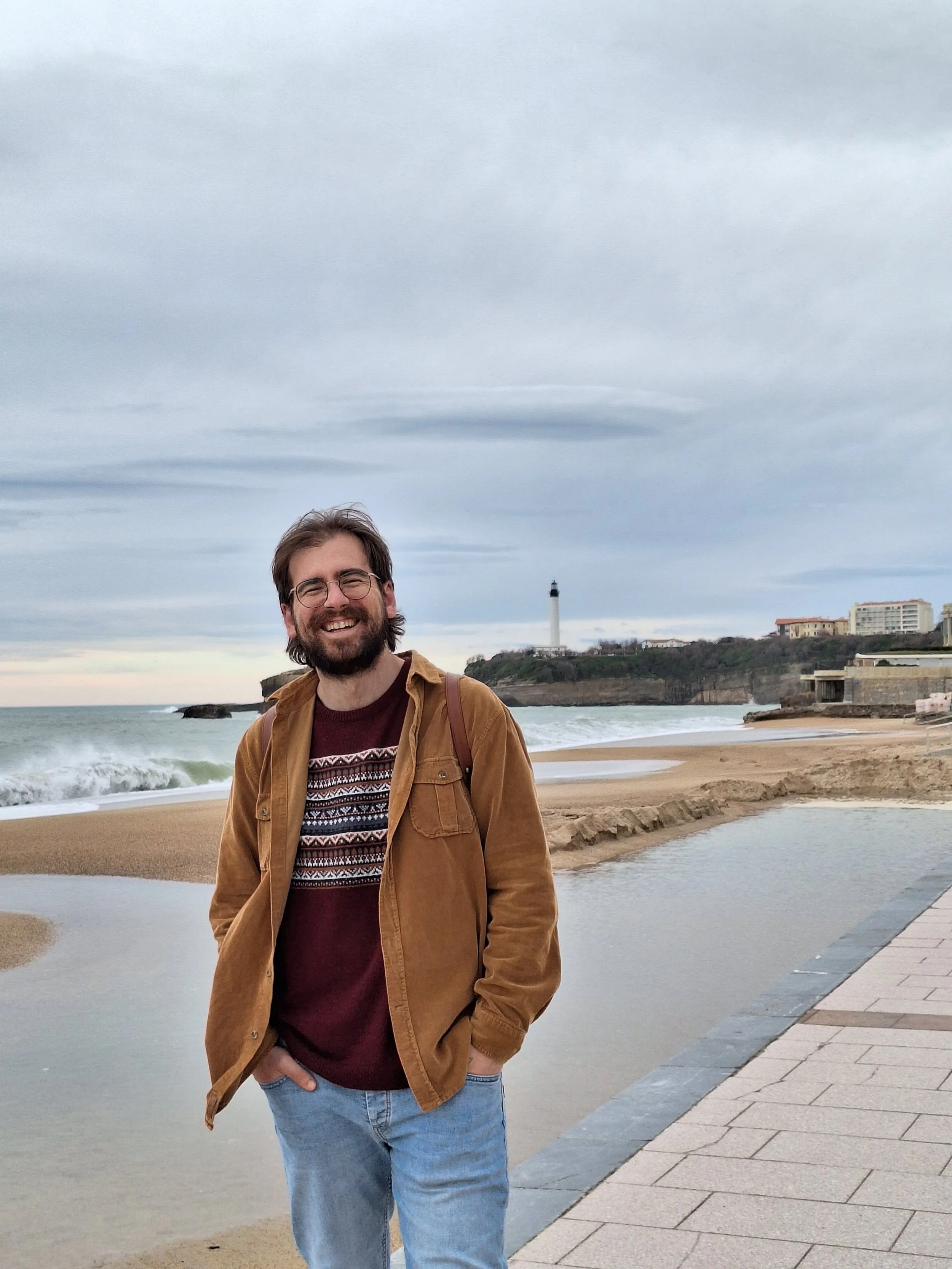 A man with glasses, beard, and long hair, wearing a brown jacket and maroon sweater, standing on a beach with a lighthouse and cliffs in the background.