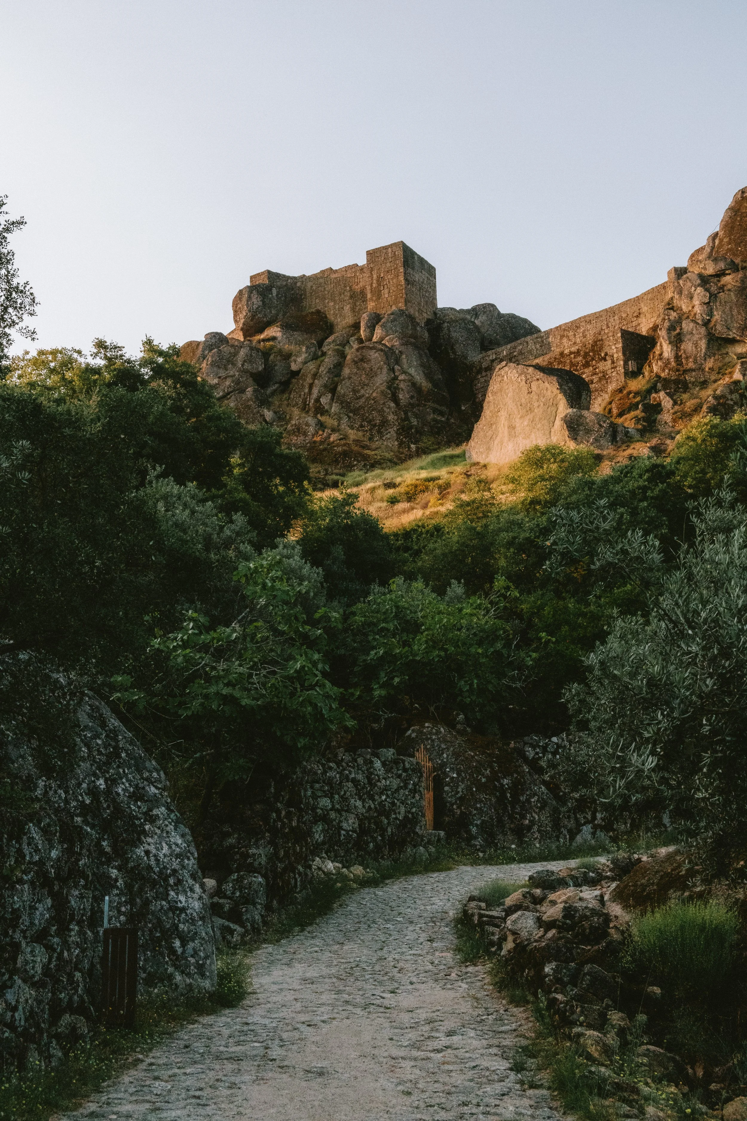 A stone pathway leading towards a rocky hill with an ancient stone fortress on top, surrounded by greenery.