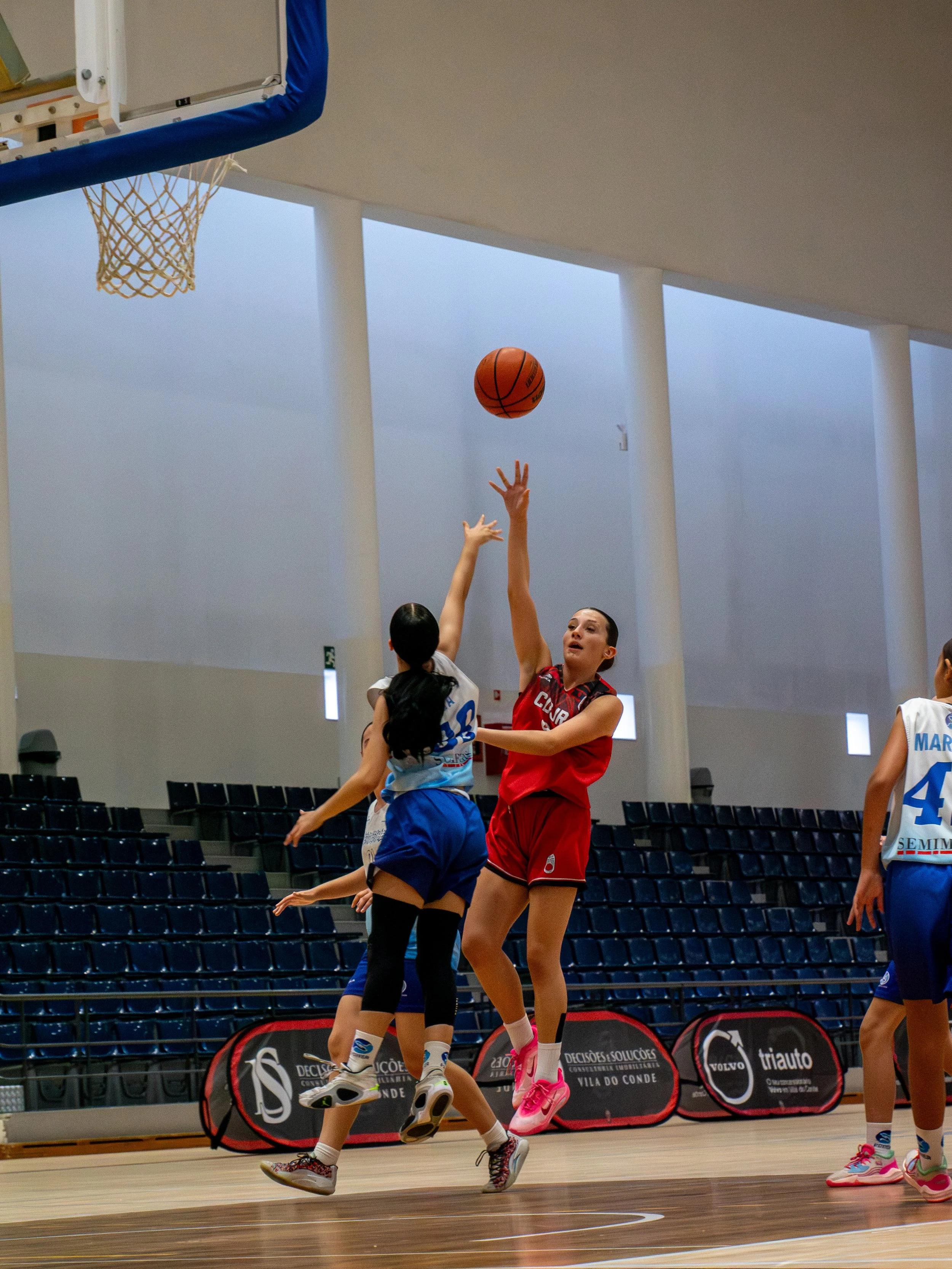 Two girls playing basketball in an indoor court, one in a red jersey jumping to shoot and the other in a blue jersey defending.