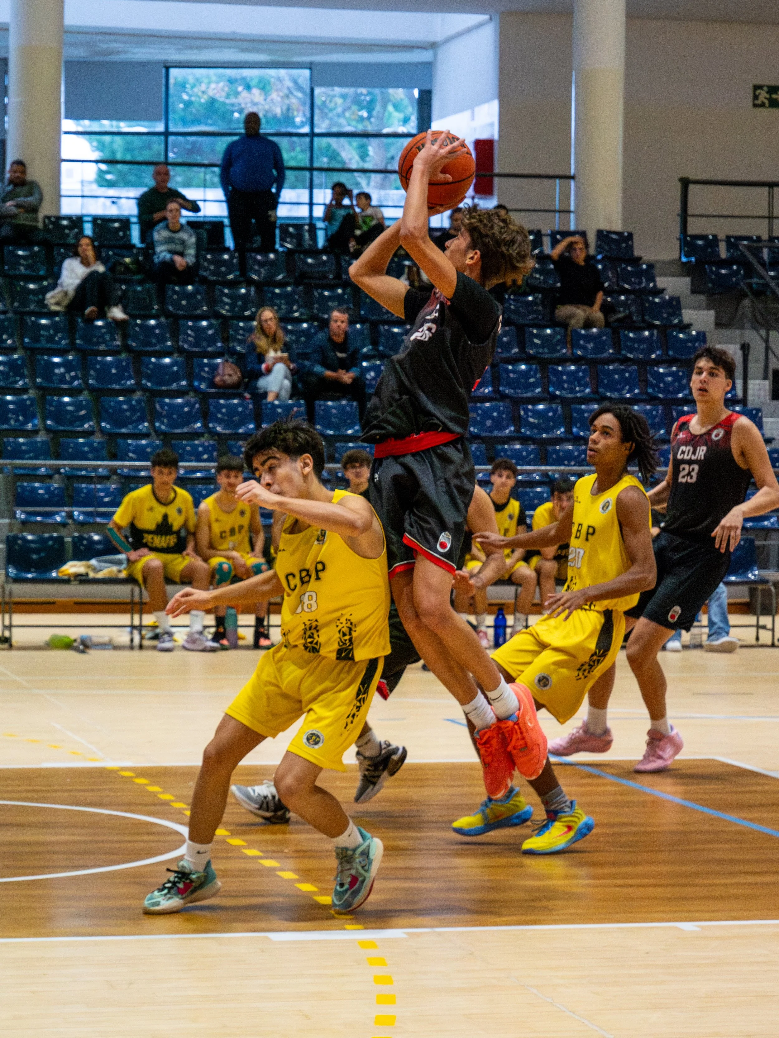 Young basketball player in black uniform jumping to shoot the ball while players in yellow uniforms attempt to block, during a youth basketball game in an indoor gym.