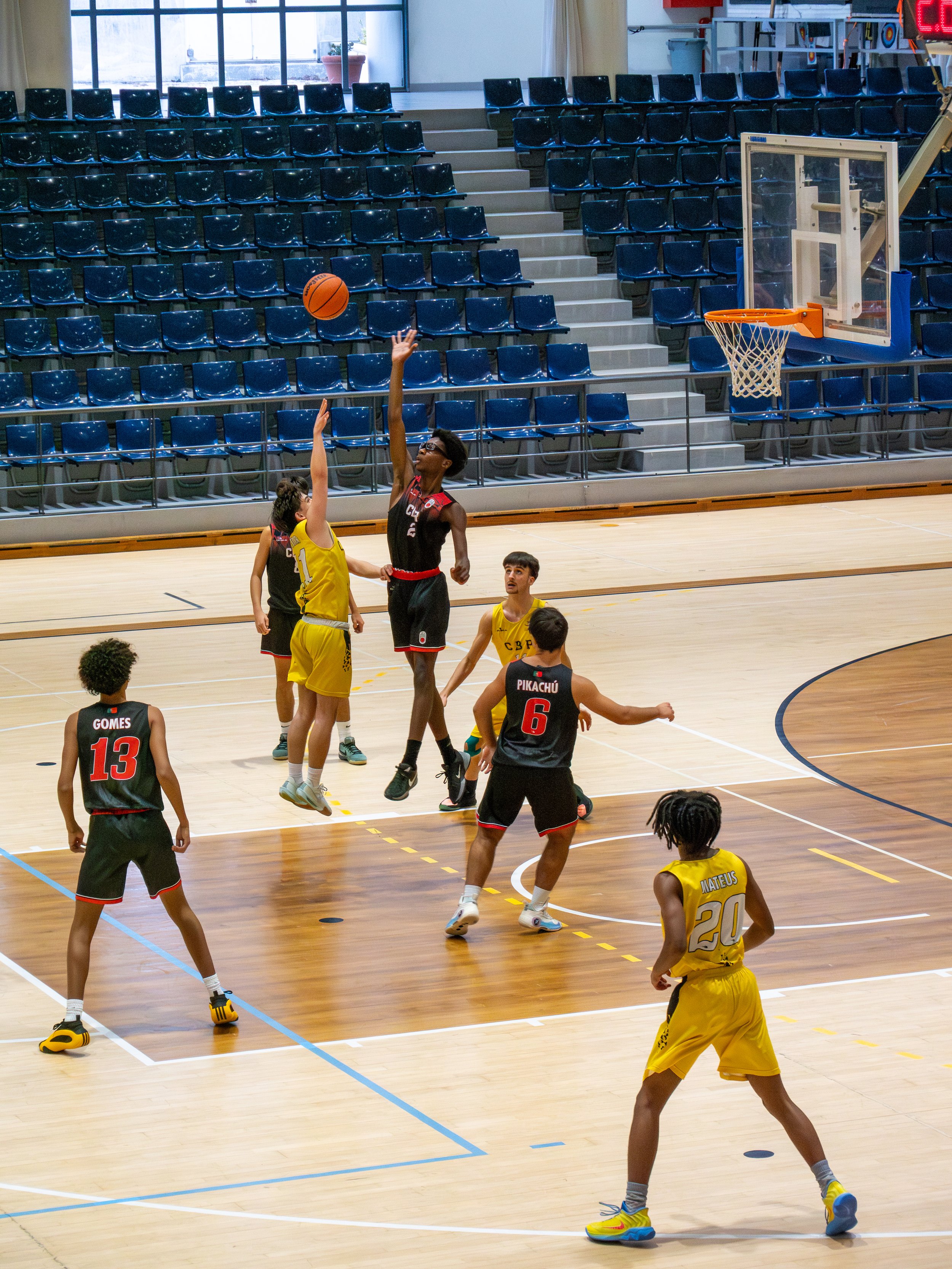 Children playing basketball on an indoor court, with one player attempting a shot while others watch and defend.
