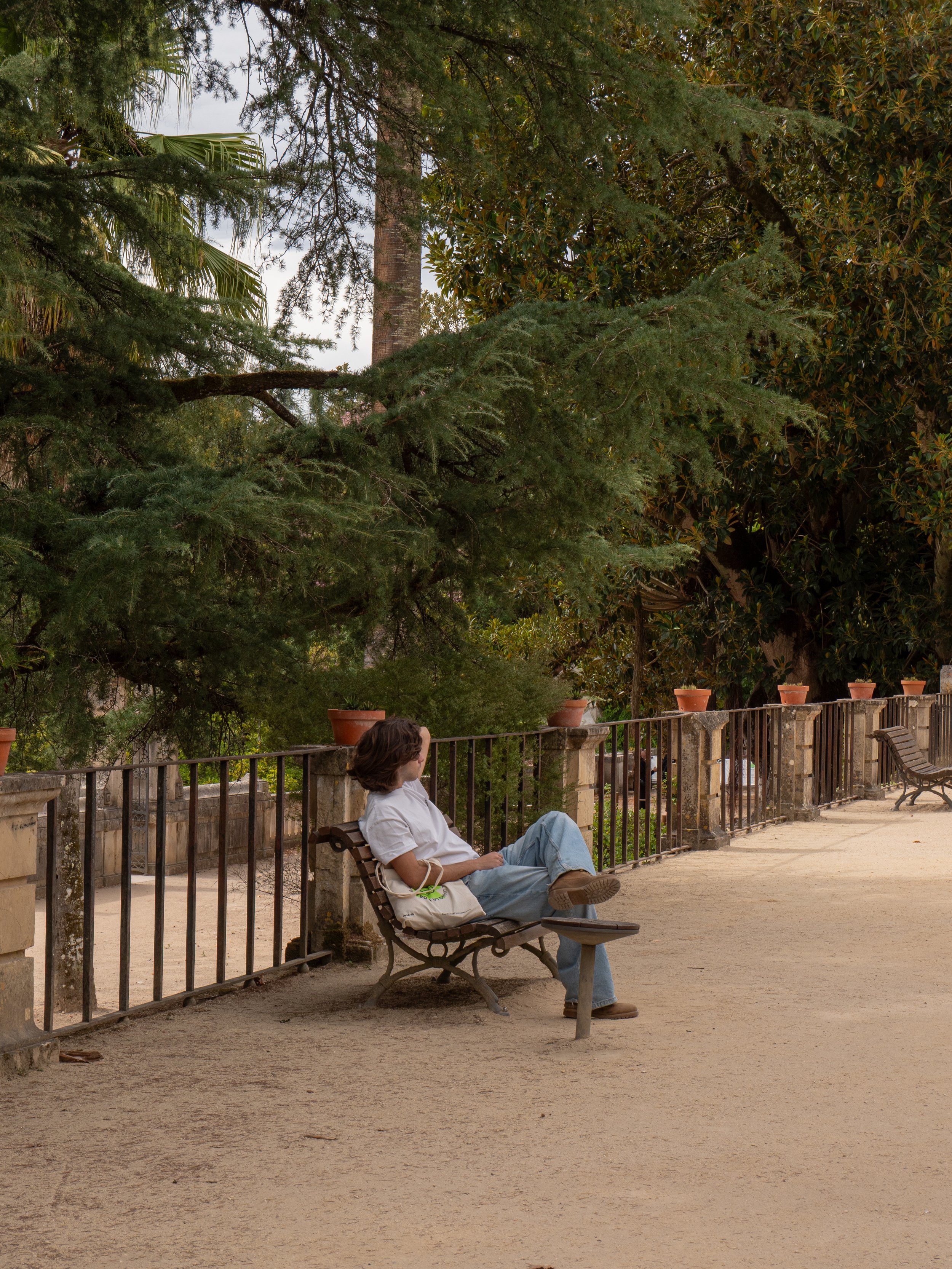 A woman with short brown hair sitting on a park bench with her legs crossed, facing to the side, surrounded by trees and potted plants on a sunny day.