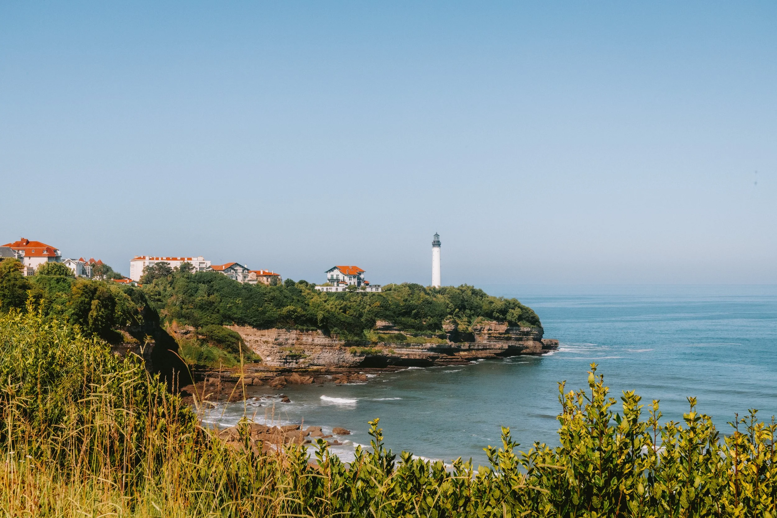 A coastal landscape featuring green hills with white buildings and red roofs, a tall lighthouse, and a calm ocean under a clear blue sky.