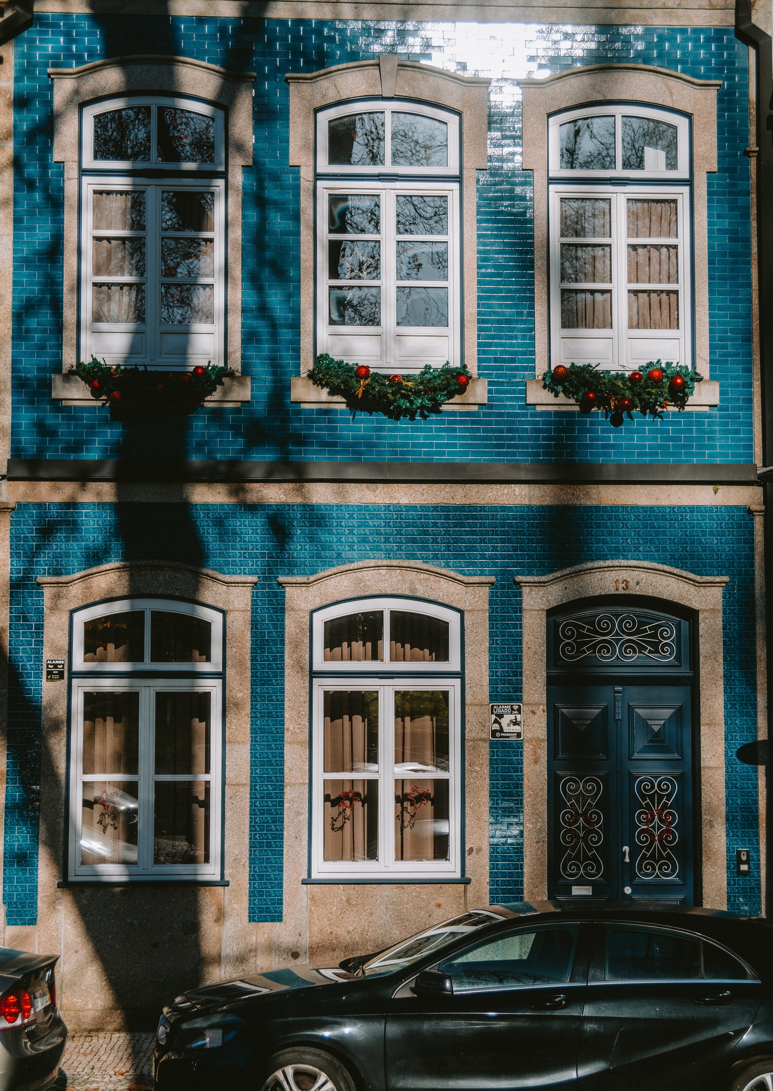 A blue multi-story building with three arched windows on both the second and first floors, decorated with Christmas garlands and red ornaments. There is a black ornate front door and parked cars in front.