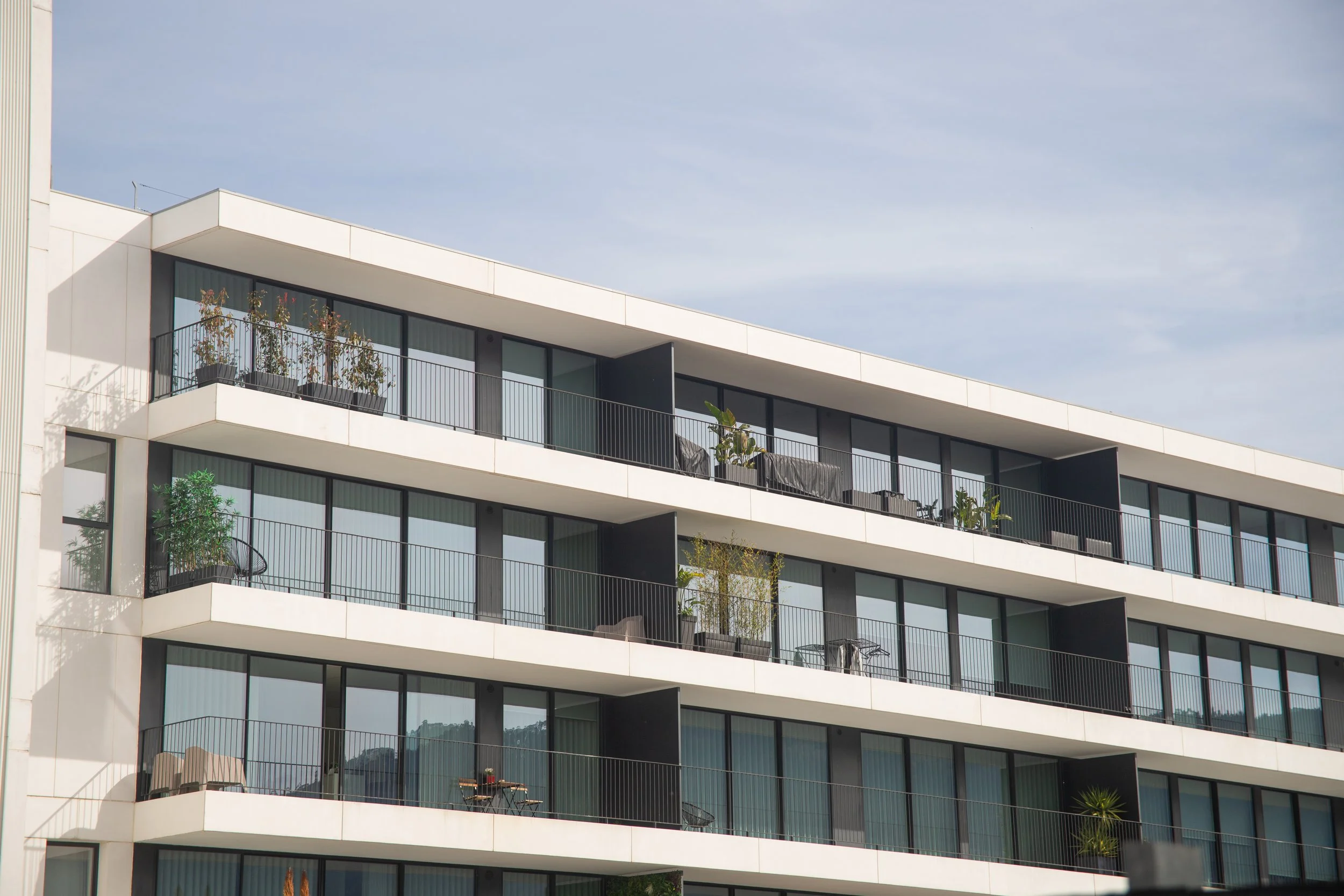 Exterior view of a modern multi-story apartment building with balconies featuring glass doors, plants, and outdoor furniture against a cloudy sky.