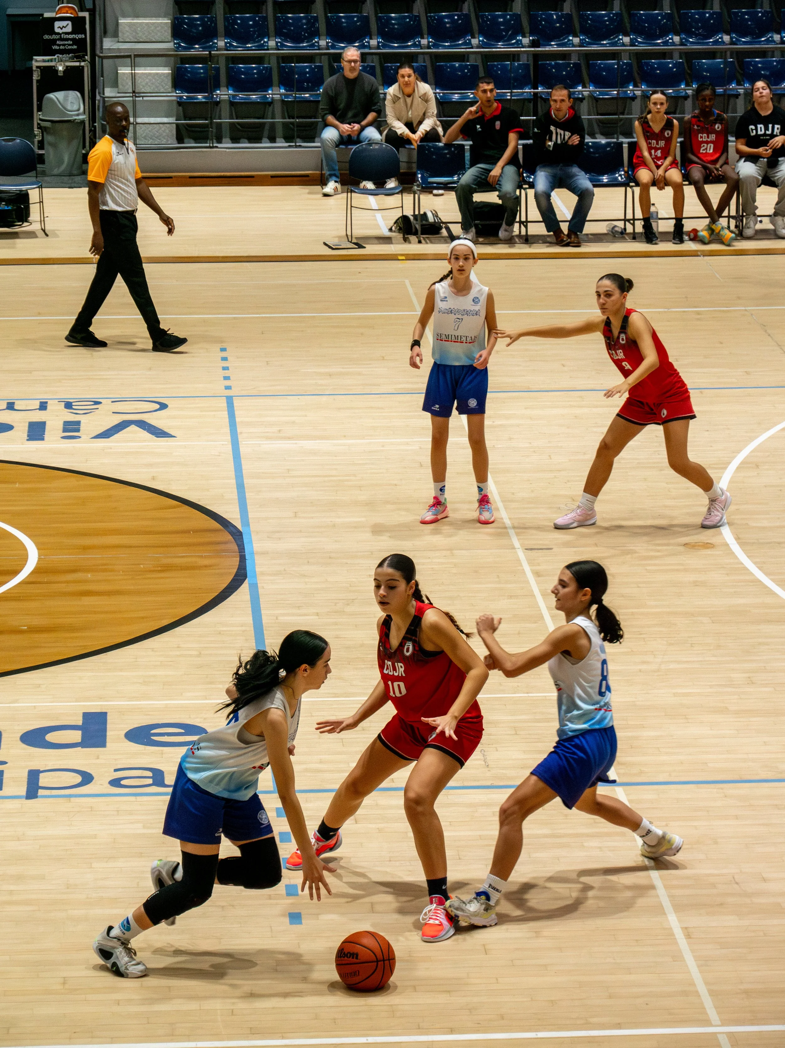 Three girls playing basketball with two defending and one controlling the ball, on a gymnasium court with spectators seated in the background.