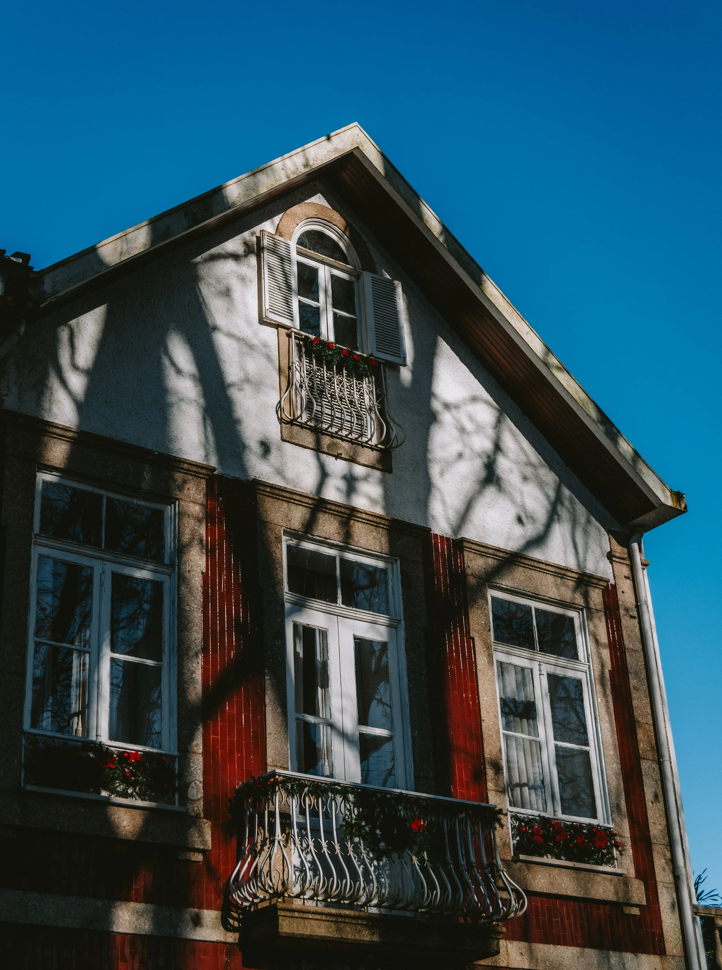A multi-story house with white and red walls, shadow of tree branches on the facade, white-framed windows, some with flower boxes, and an open window with shutters on the upper floor. The house is under a clear blue sky.