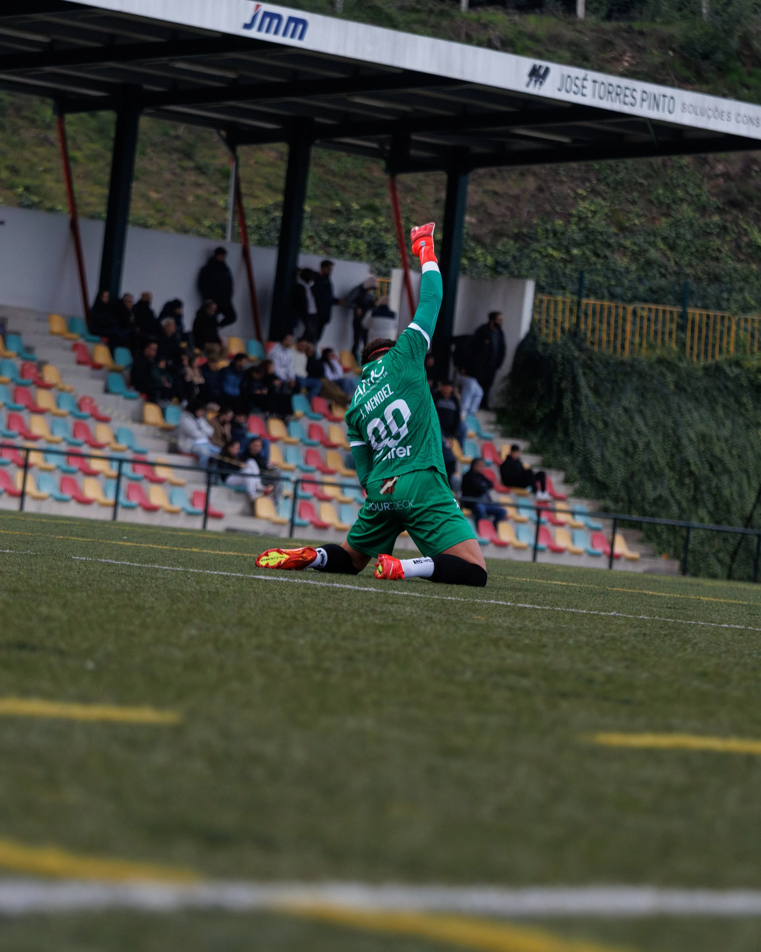 A soccer player in a green uniform kneeling and pointing upward on a field, with spectators in the background.