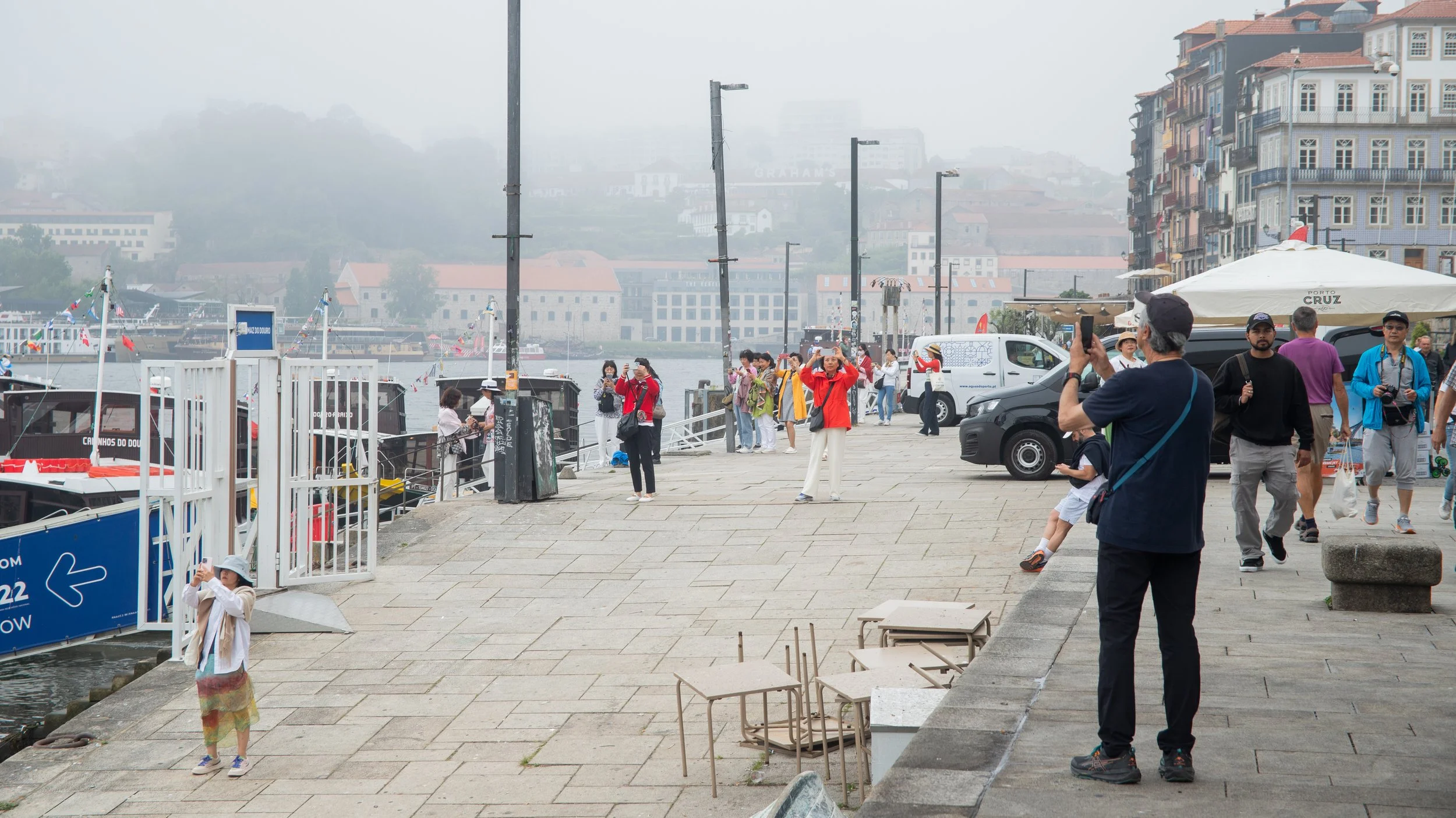People taking photos near the harbor in foggy weather, with boats docked and buildings in the background.