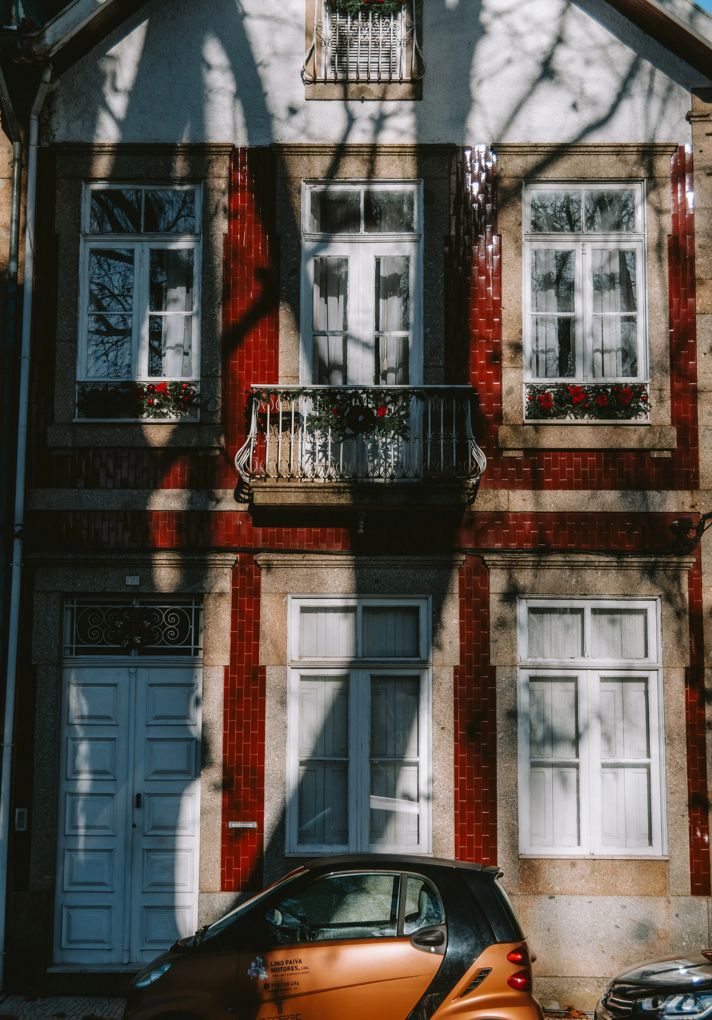 A multi-story residential building with white-framed windows, some with flower boxes, a small balcony with decorative railing, and red tile accents on the facade. A small car is parked in front.