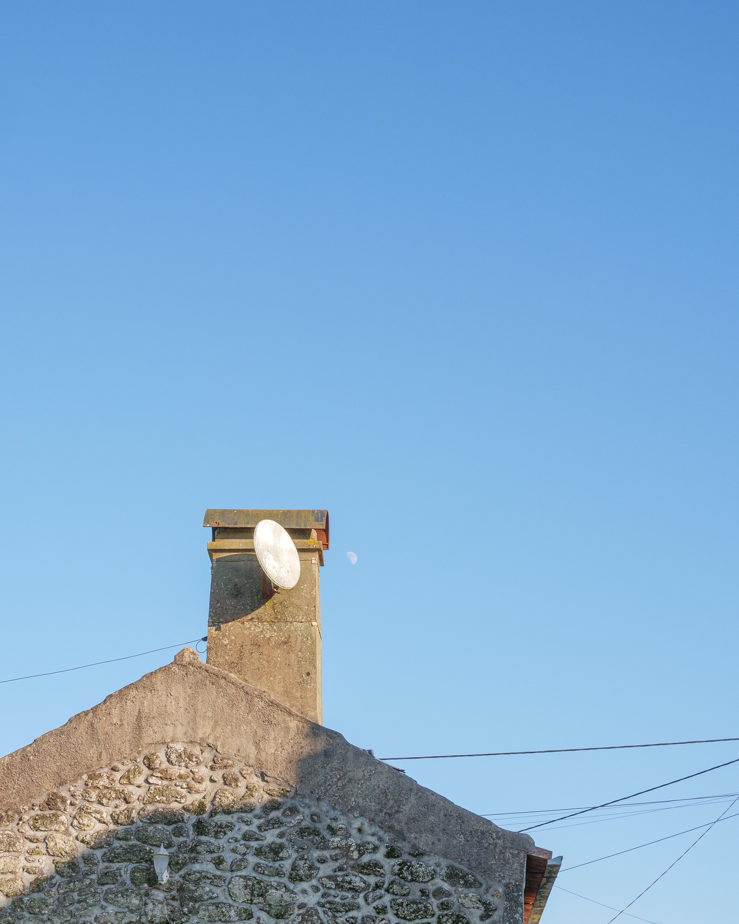 View of a house chimney with a satellite dish, with the moon visible in the sky to the right of the chimney and electrical wires crossing the sky.